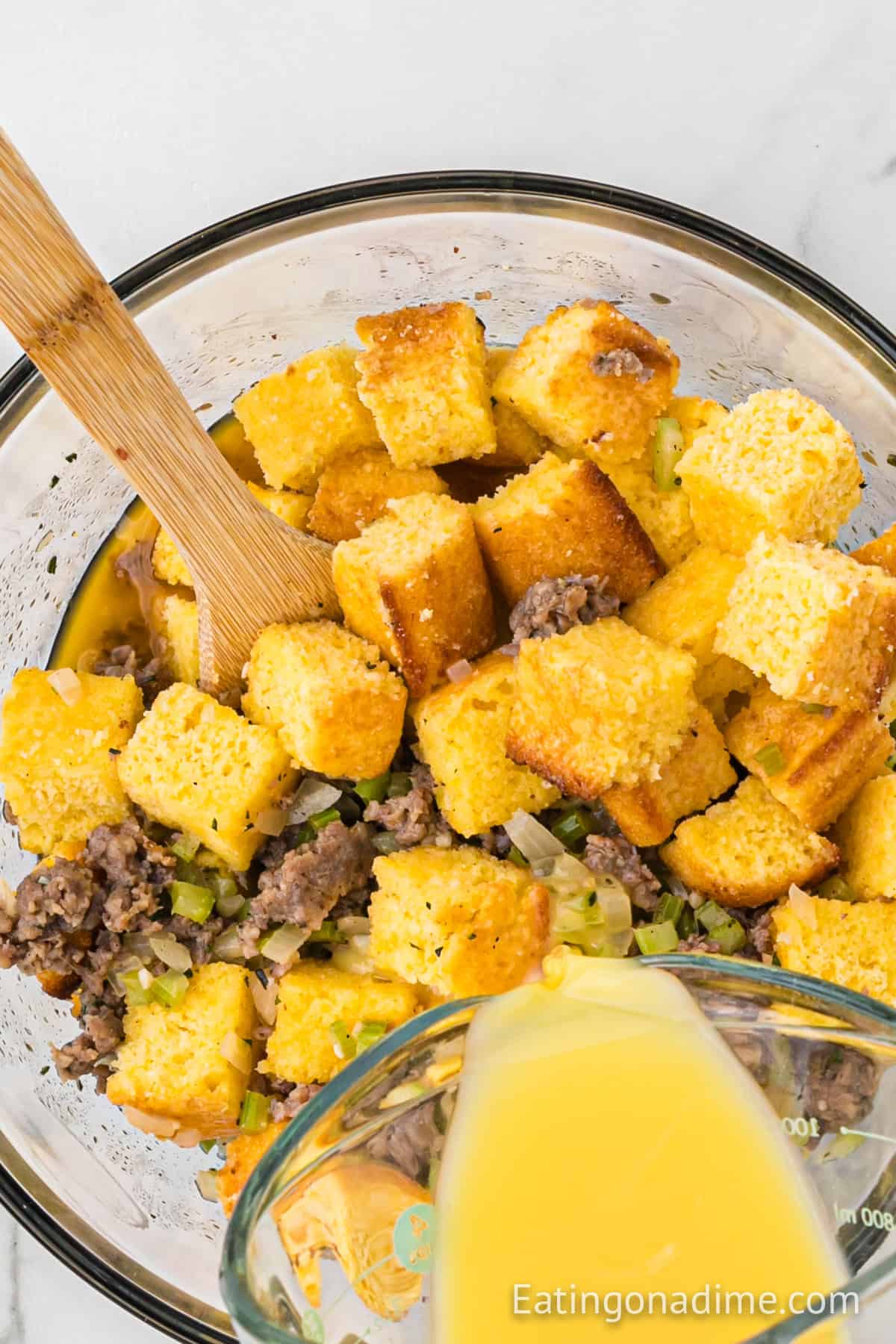 A bowl with cubed cornbread, cooked sausage, and diced celery sits ready for transformation into a savory Cornbread Sausage Stuffing recipe. A wooden spoon rests in the bowl as a pitcher pours liquid over this rich mixture. The image is courtesy of Eating on a Dime.