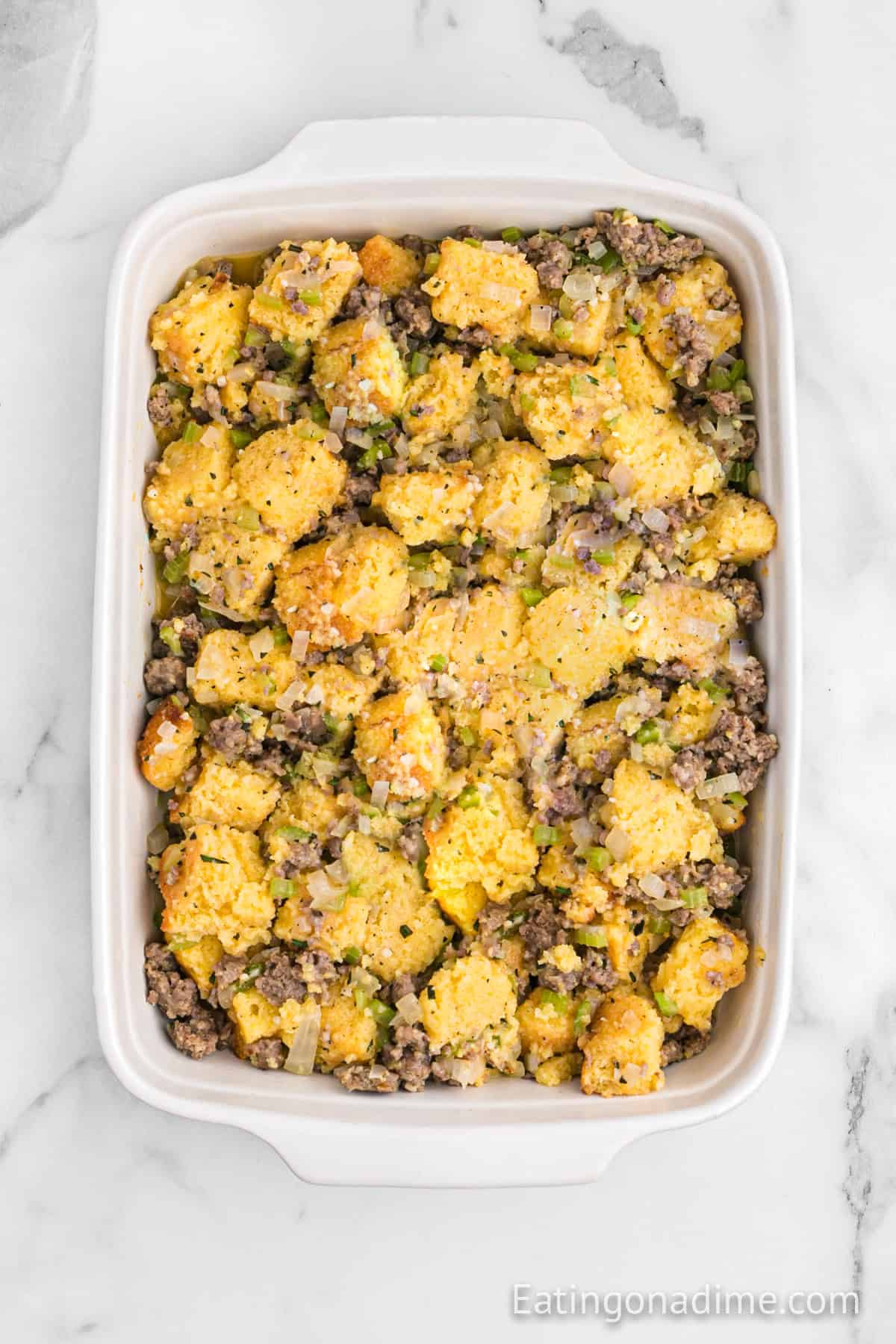 A rectangular dish showcases a cornbread sausage stuffing recipe, richly mixed with ground meat and chopped vegetables, including onions and celery, elegantly placed on a marble surface.