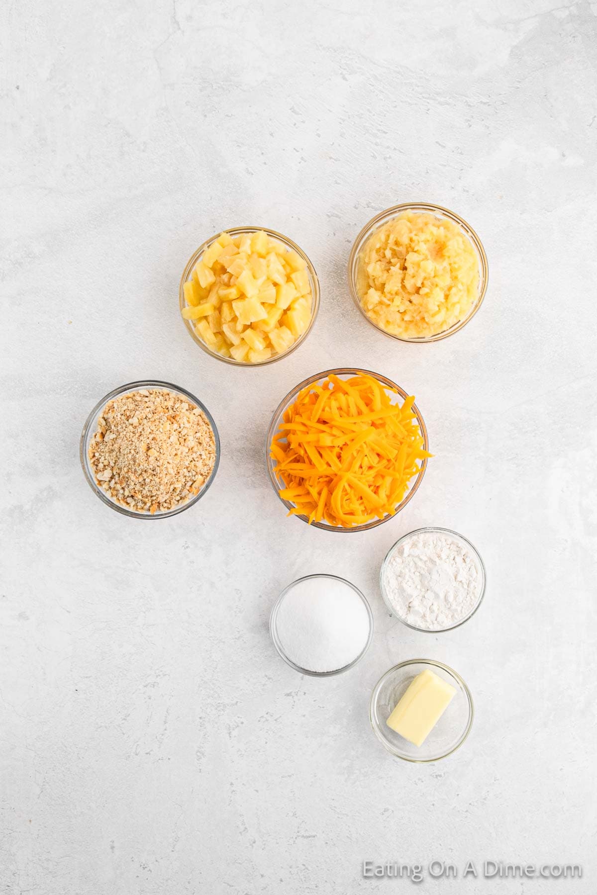 Top view of food ingredients arranged in small bowls on a light surface. The ingredients for a delightful pineapple casserole include chopped potatoes, mashed potatoes, shredded cheddar cheese, breadcrumbs, flour, sugar, and a small piece of butter.