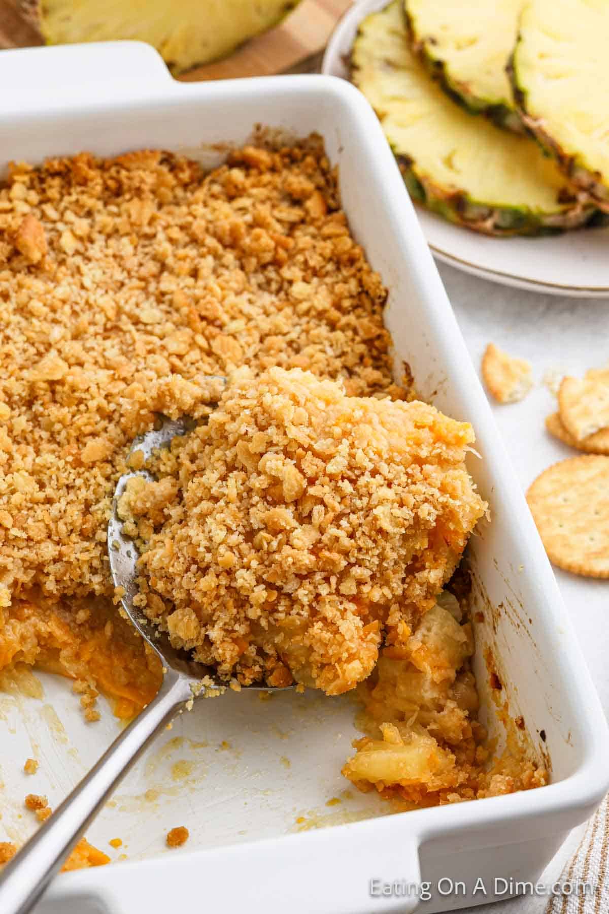 A white baking dish filled with a golden-brown Pineapple Casserole topped with crumbly cracker crust. A serving spoon is lifting a portion, displaying the mixture of crushed pineapple and crunchy topping. A plate with pineapple slices is in the background.
