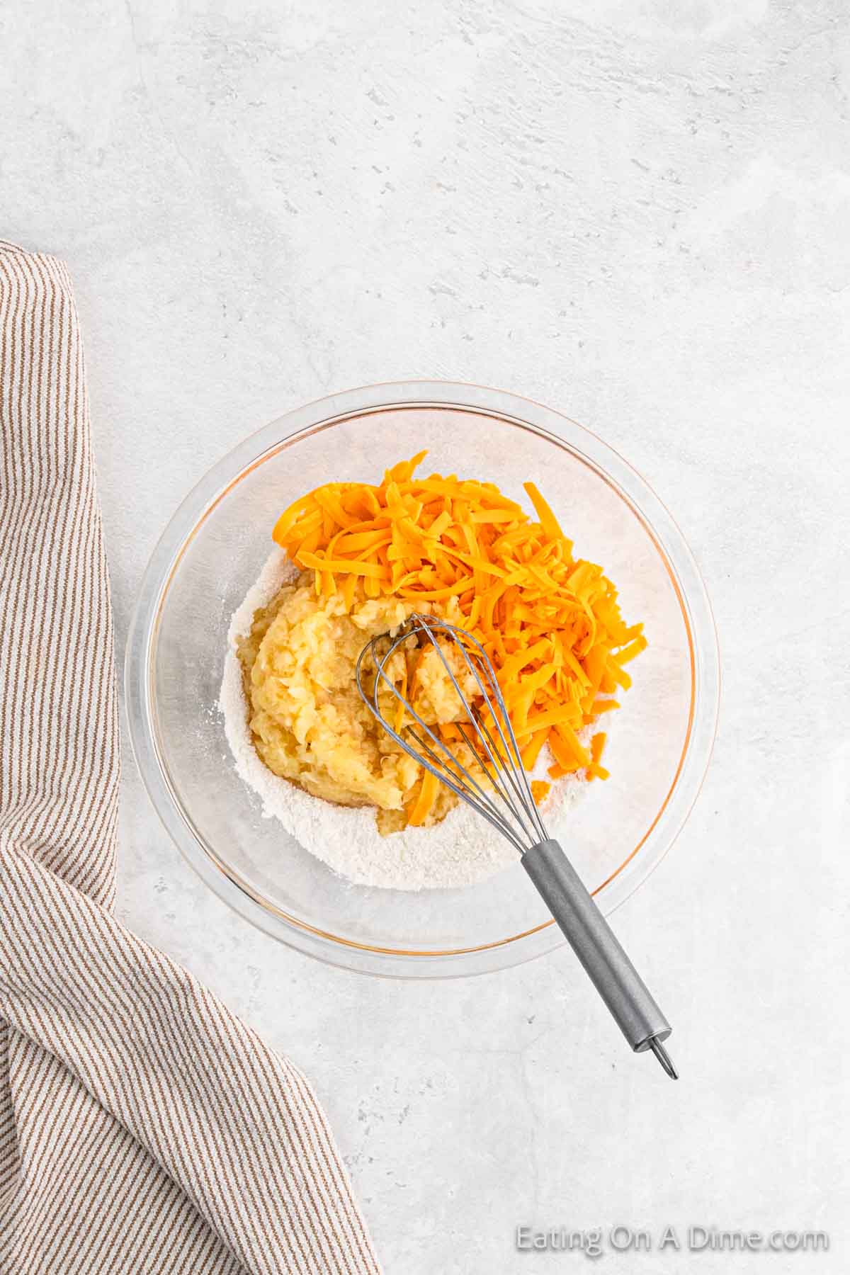 A glass mixing bowl contains shredded cheddar cheese on top of a flour and mashed potato mixture. A whisk rests in the bowl, ready to mix the ingredients for a delicious Pineapple Casserole. A beige striped kitchen towel is placed beside the bowl on a textured white surface.