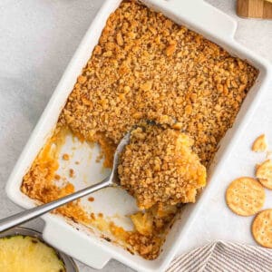 A rectangular baking dish contains a partially served pineapple casserole with a golden-brown, crumbly topping. A serving spoon rests inside the dish. Crackers partially crumble on the right, and a small piece of pineapple is visible in the bottom left corner.