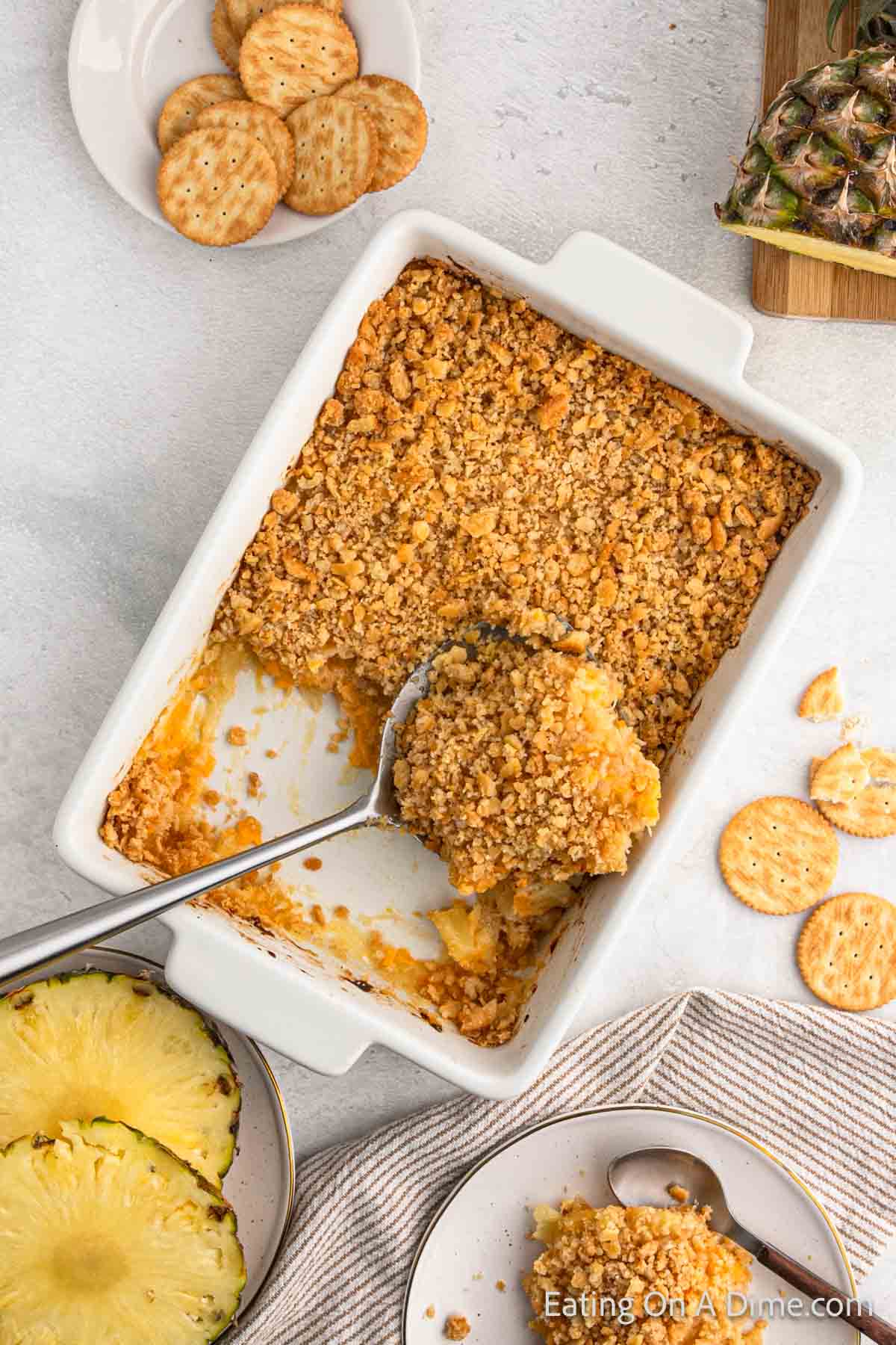 A white rectangular baking dish contains a partially served Pineapple Casserole with a crumb topping. A serving spoon rests inside the dish. Nearby, there are round crackers, fresh pineapple slices on plates, and a small cutting board with additional pineapple.