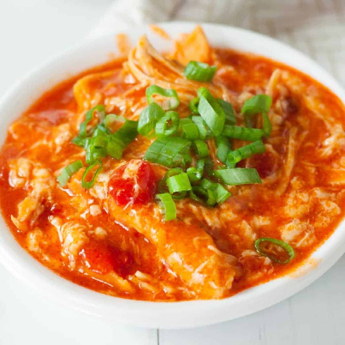 A bowl of creamy buffalo chicken dip, reminiscent of a crock pot buffalo chicken enchilada casserole, is topped with sliced green onions. The rich orangish-red color and chunks of chicken and tomatoes lend a hearty, appetizing appearance.