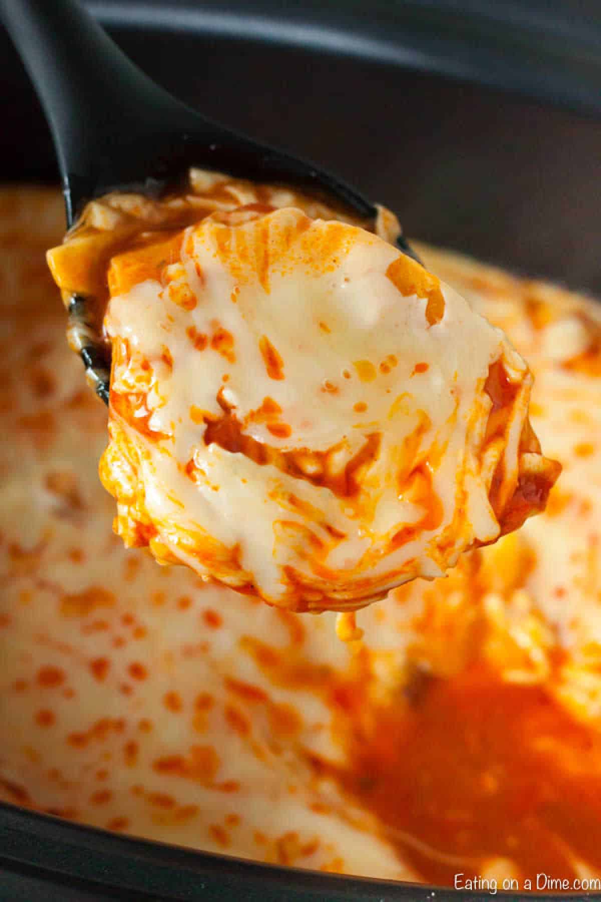 A close-up of a spoon scooping creamy buffalo chicken dip from a crock pot. The dip is orange with melted cheese on top, reminiscent of a gooey enchilada casserole.