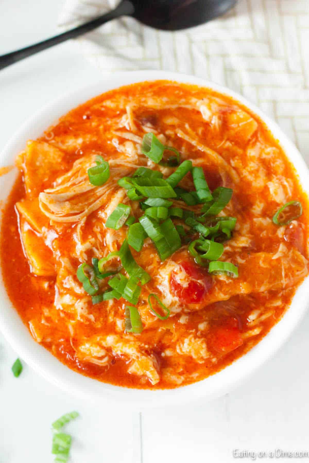 A bowl of chicken tortilla soup, reminiscent of a crock pot buffalo chicken creation, is topped with chopped green onions. The soup's rich red color envelops shredded chicken, tomatoes, and tortilla pieces. A spoon rests on a white napkin in the background.