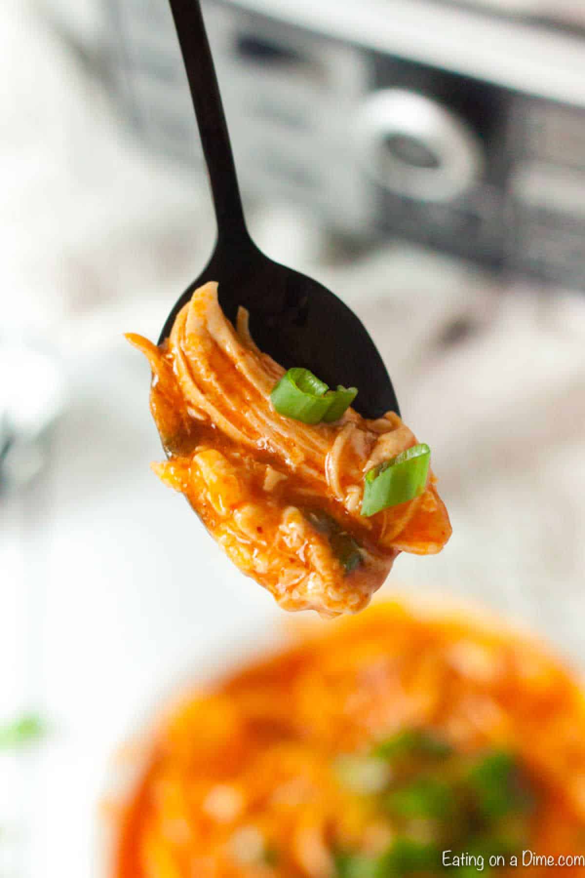 A black spoon holds a portion of crock pot buffalo chicken enchilada casserole, drenched in a rich red sauce and garnished with green onions. A bowl of this savory dish is blurred in the background.