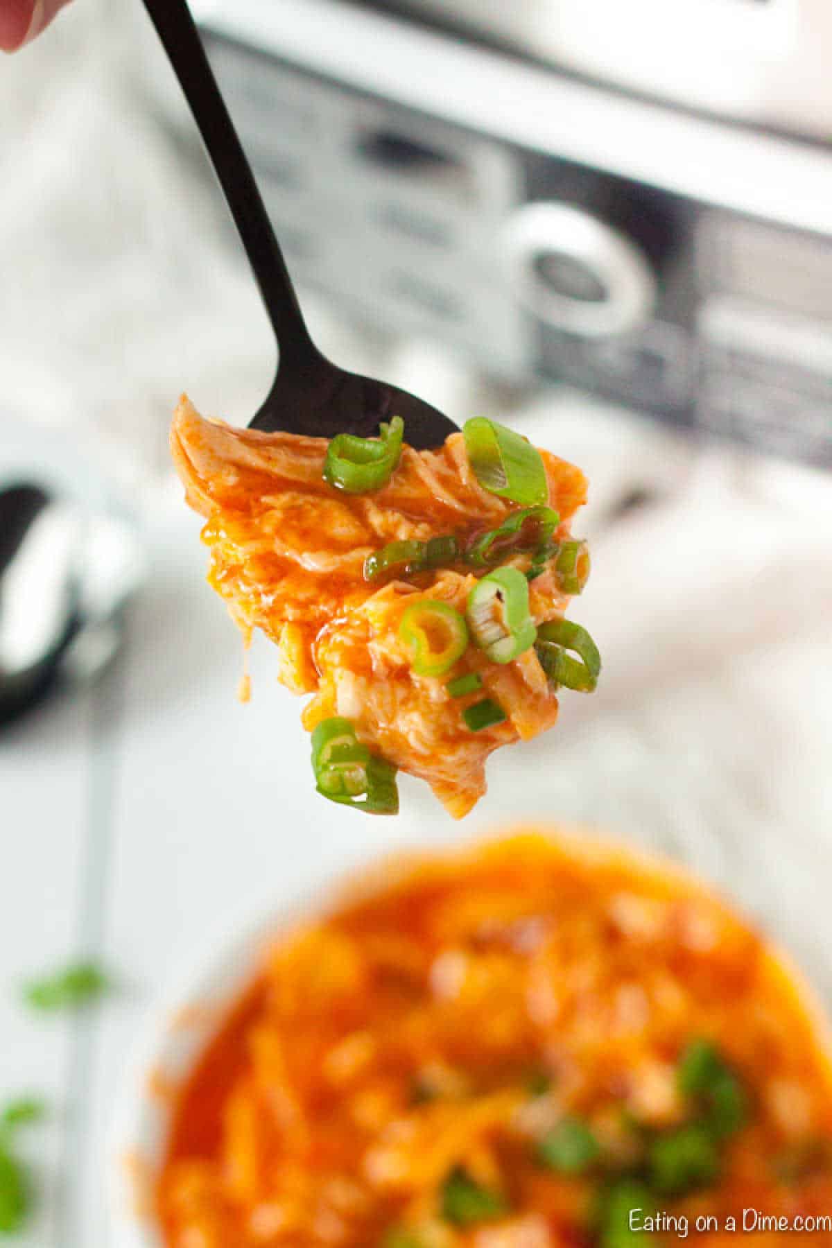 A fork holds a portion of orange-colored buffalo chicken dip, topped with sliced green onions. In the background, more of this crock pot creation can be seen in a white bowl, along with a blurred kitchen appliance.