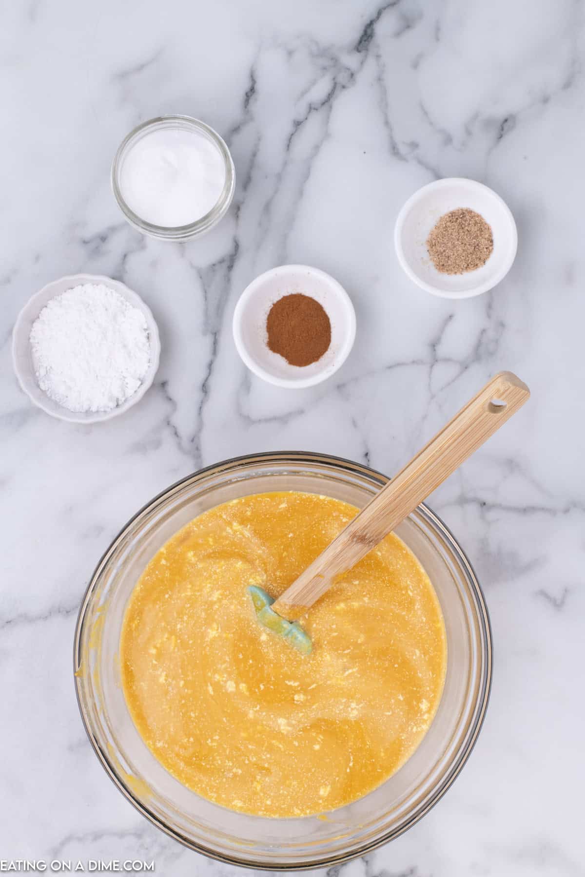 A glass bowl filled with a yellow batter mixture and a spatula on a marble countertop. Surrounding the bowl are small dishes containing various powdered ingredients like sugar, flour, cinnamon, and another brown powdered spice perfect for crafting butternut squash pie.