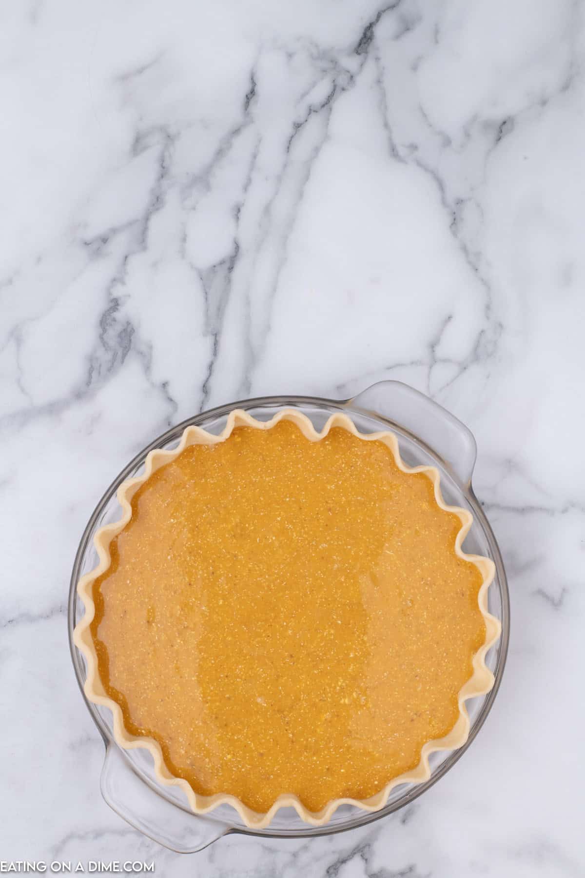 A pumpkin pie with a crimped edge sits in a glass pie dish on a white marble countertop. The unbaked butternut squash pie filling is smooth and rich, boasting a vibrant orange hue. The dish handles are visible on either side of the pie.