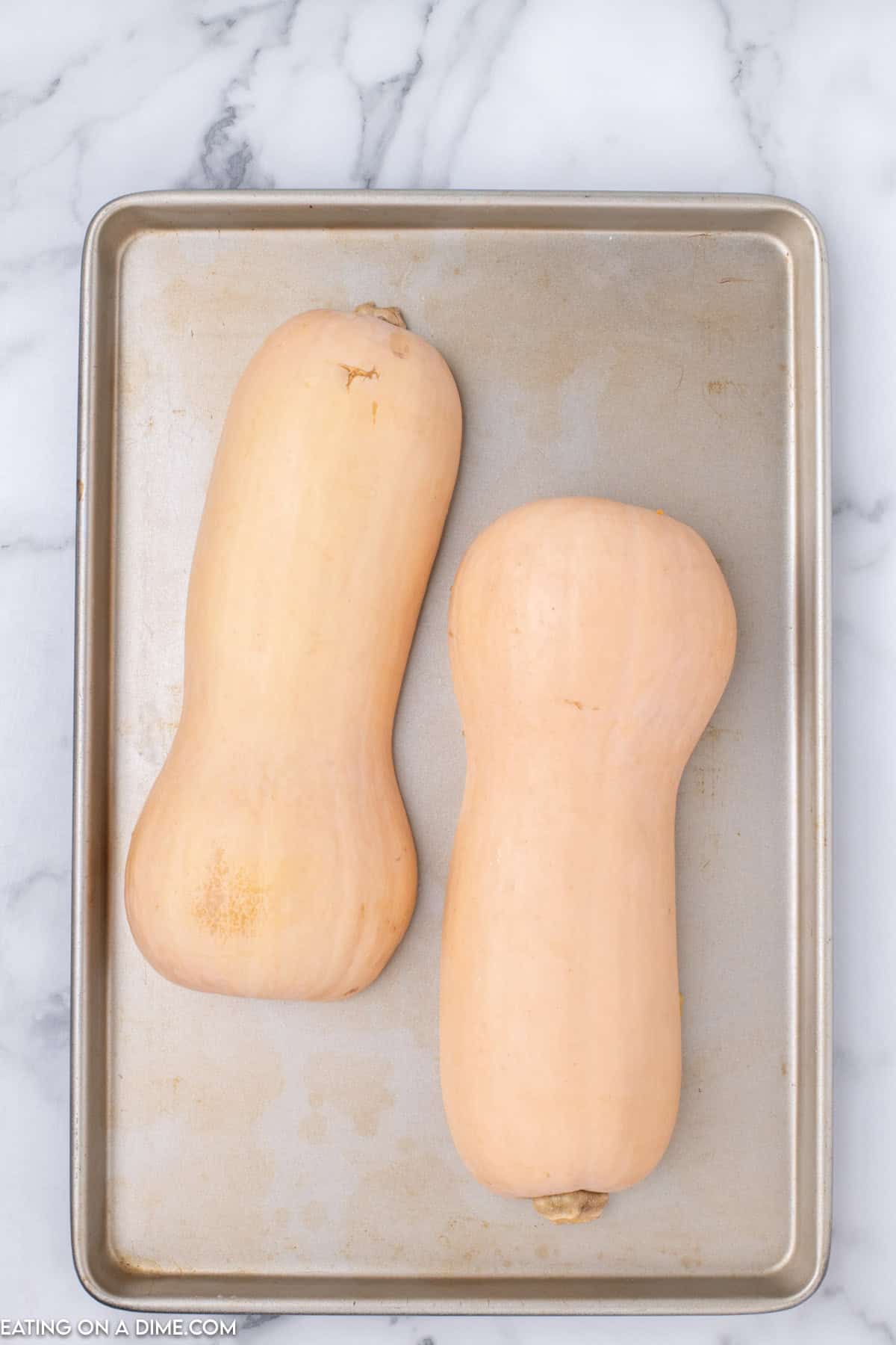 Two whole butternut squashes are placed on a baking tray, ready to be transformed into a delicious Butternut Squash Pie. The image background features a marble surface. The squashes have smooth, beige skin and are positioned side by side.