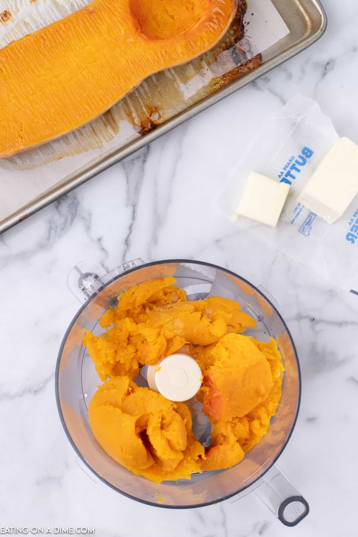 A top-down view of roasted butternut squash being blended in a food processor on a marble countertop. Nearby, there is a baking sheet with a scooped-out squash half, and an opened package of butter with a few pieces cut off, preparing for the perfect Butternut Squash Pie.