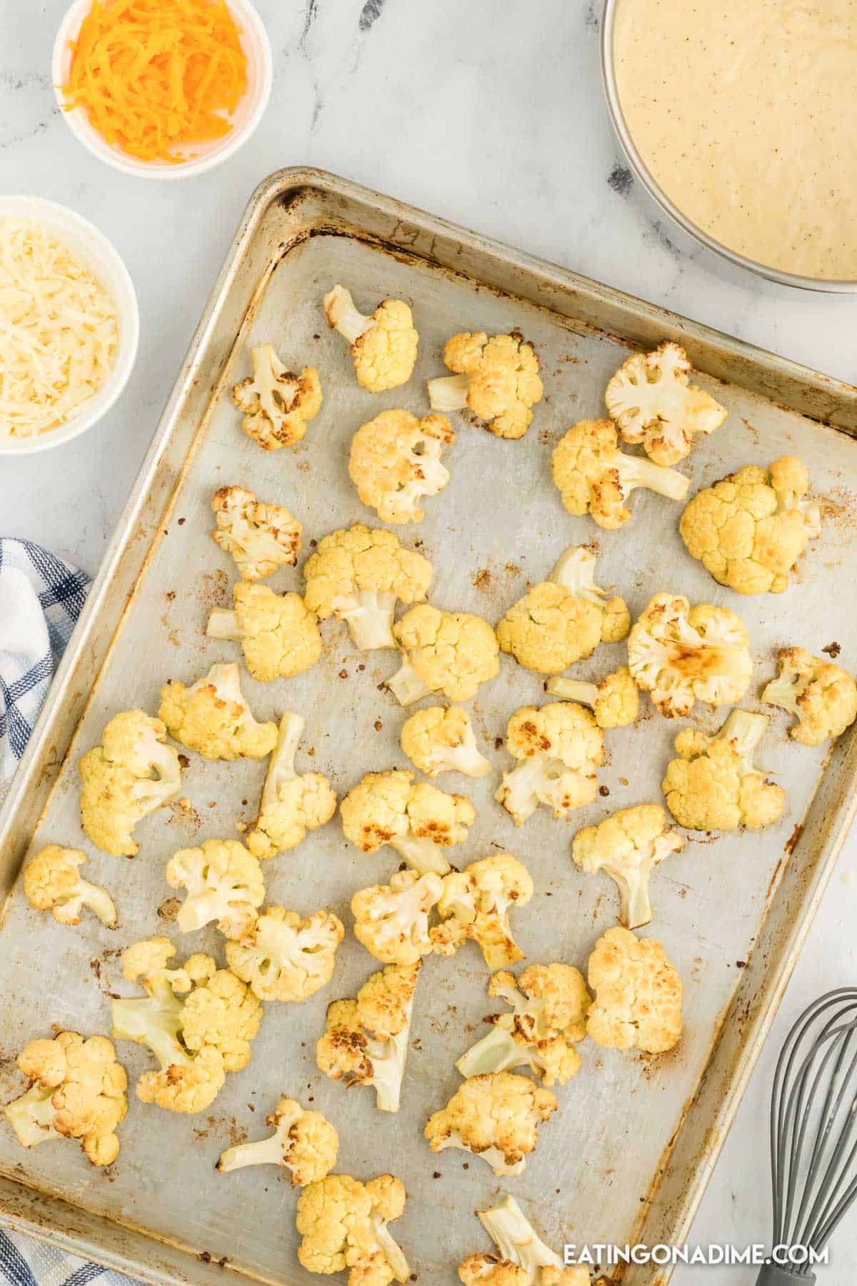 Cauliflower florets on a baking sheet with small bowls of shredded cheese 