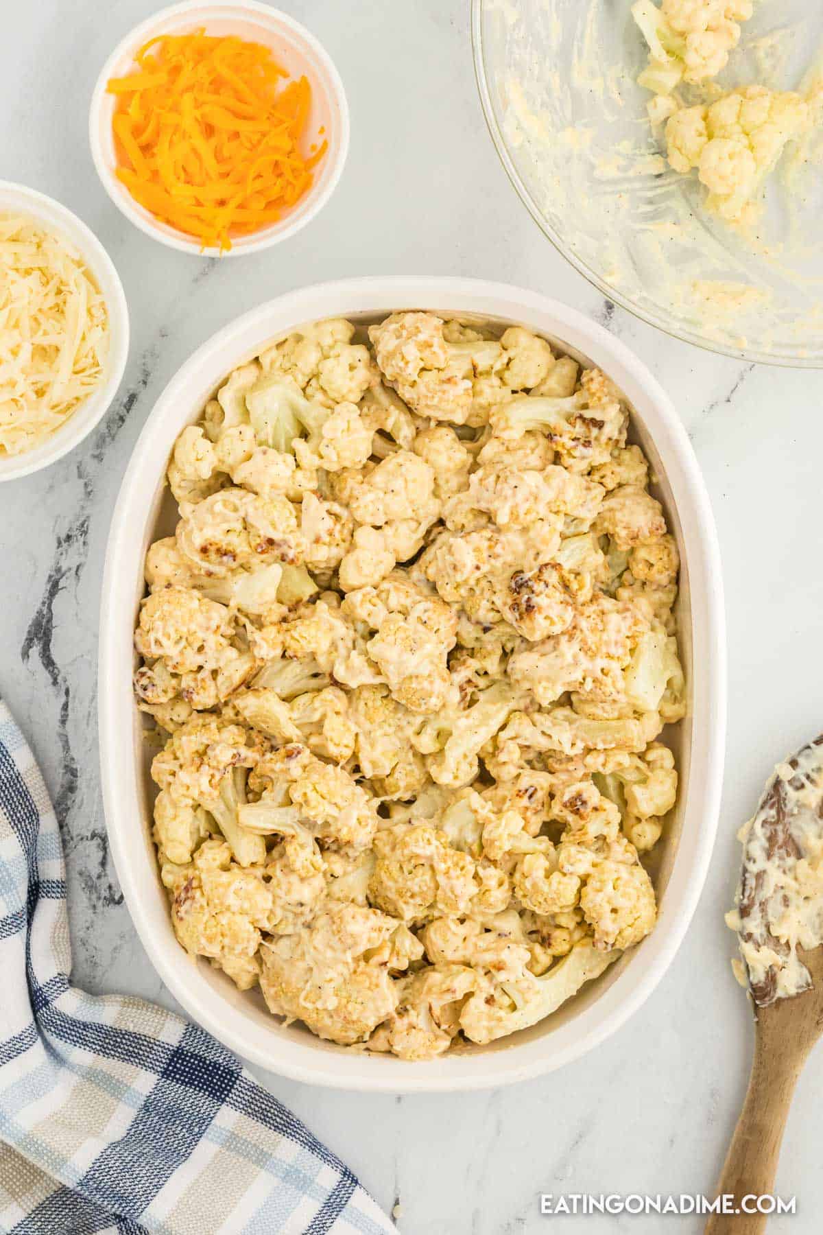 Cauliflower florets coated with the melted butter mixture in a casserole dish with small bowls of shredded cheese