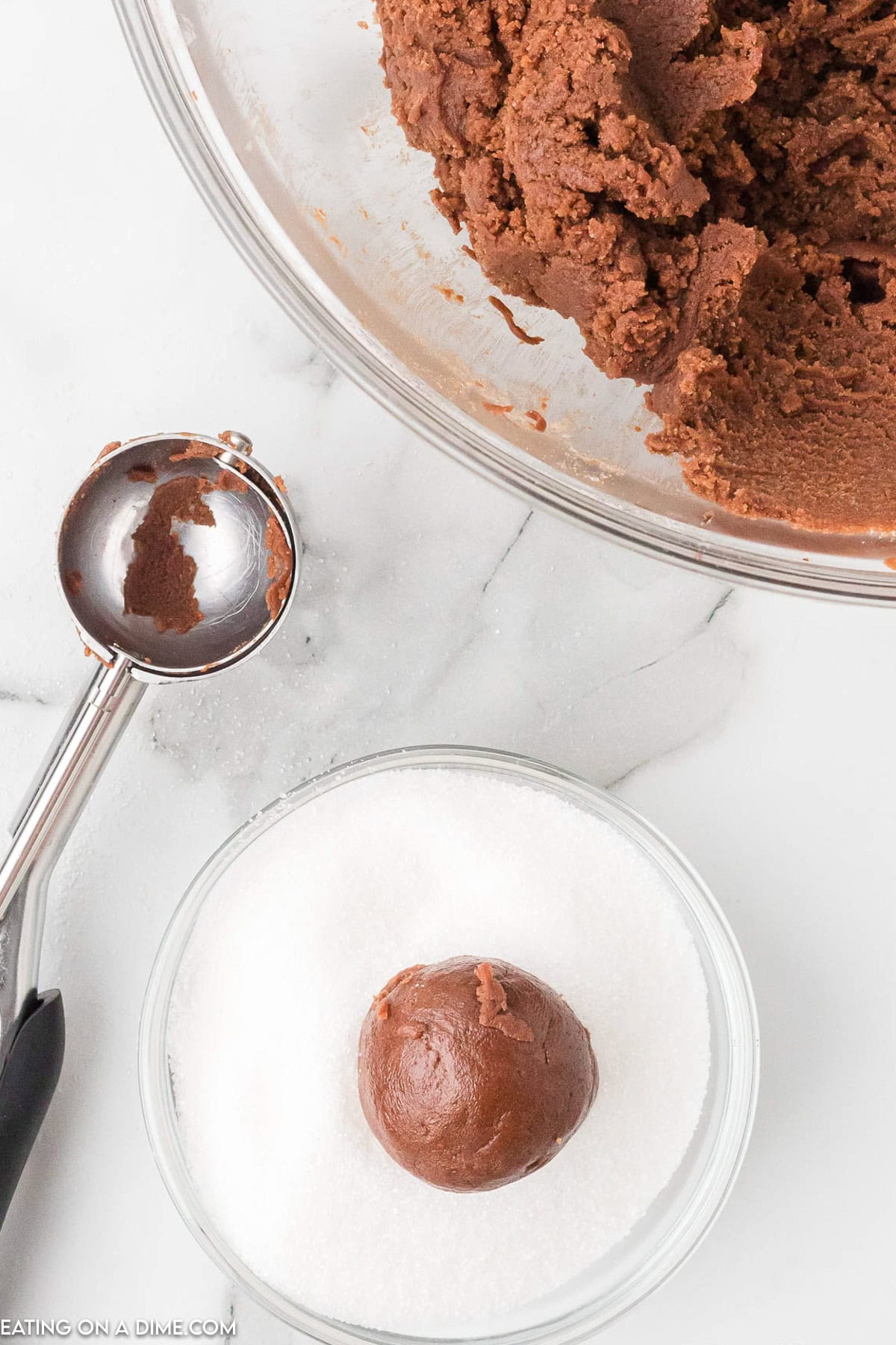 A glass bowl filled with chocolate cookie dough is on a marble countertop. Beside it, a cookie scoop with dough sits next to a smaller bowl of granulated sugar, where a round ball of chocolate dough for Chocolate Kiss Cookies is being rolled in the sugar.