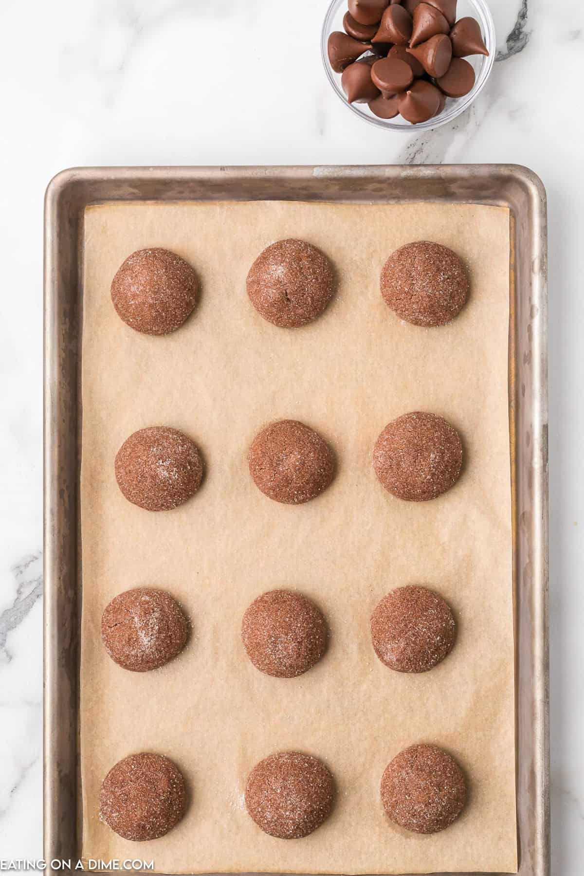 A baking sheet lined with parchment paper holds 12 round, unbaked Chocolate Kiss Cookie dough balls dusted with sugar. A small, clear bowl filled with chocolate kisses is placed beside the baking sheet on a marble countertop.