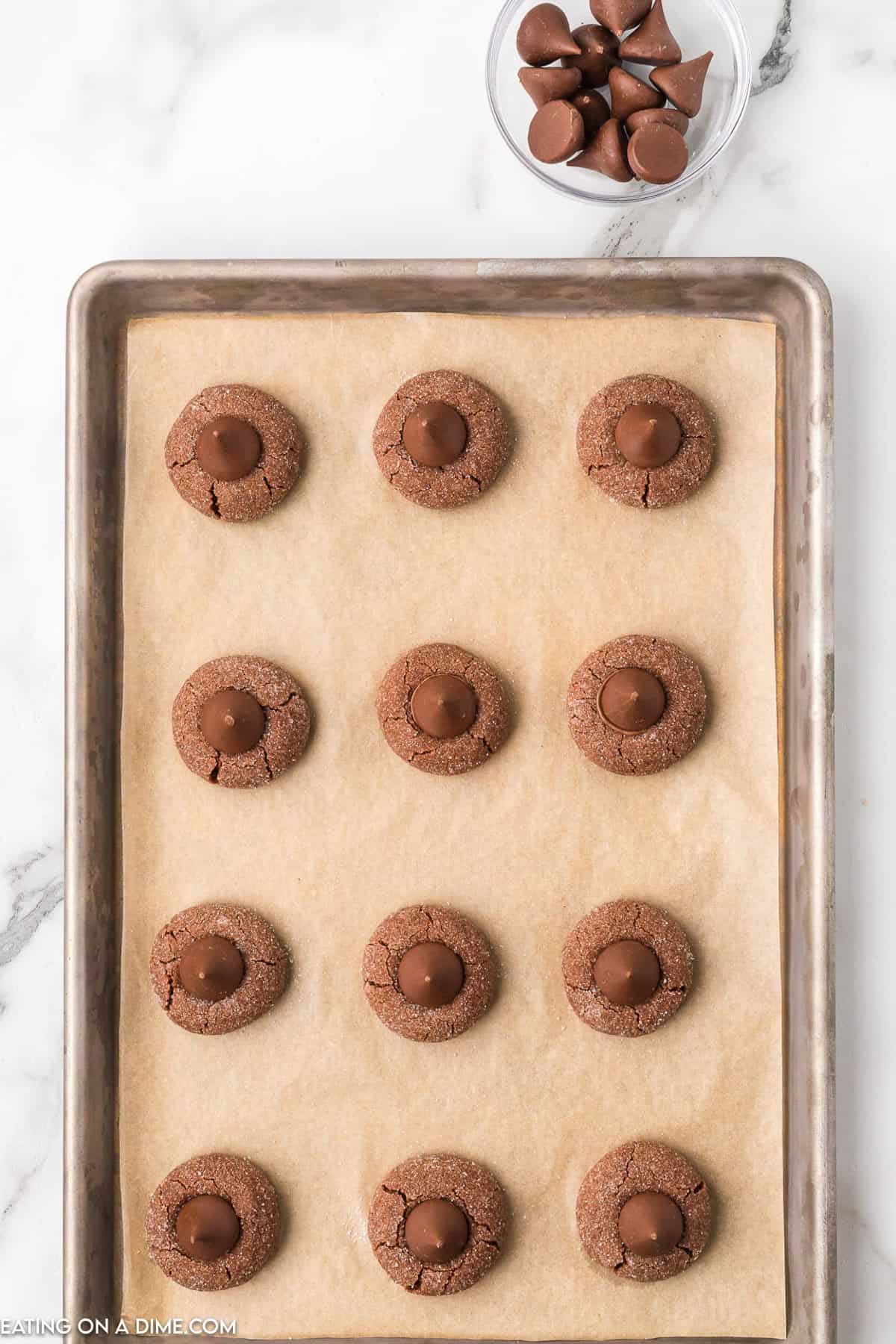 A baking tray filled with twelve round Chocolate Kiss Cookies, each topped with a Hershey's Kiss, placed on parchment paper. Nearby, a small bowl holds additional Hershey's Kisses. The tray rests on a white marble surface.