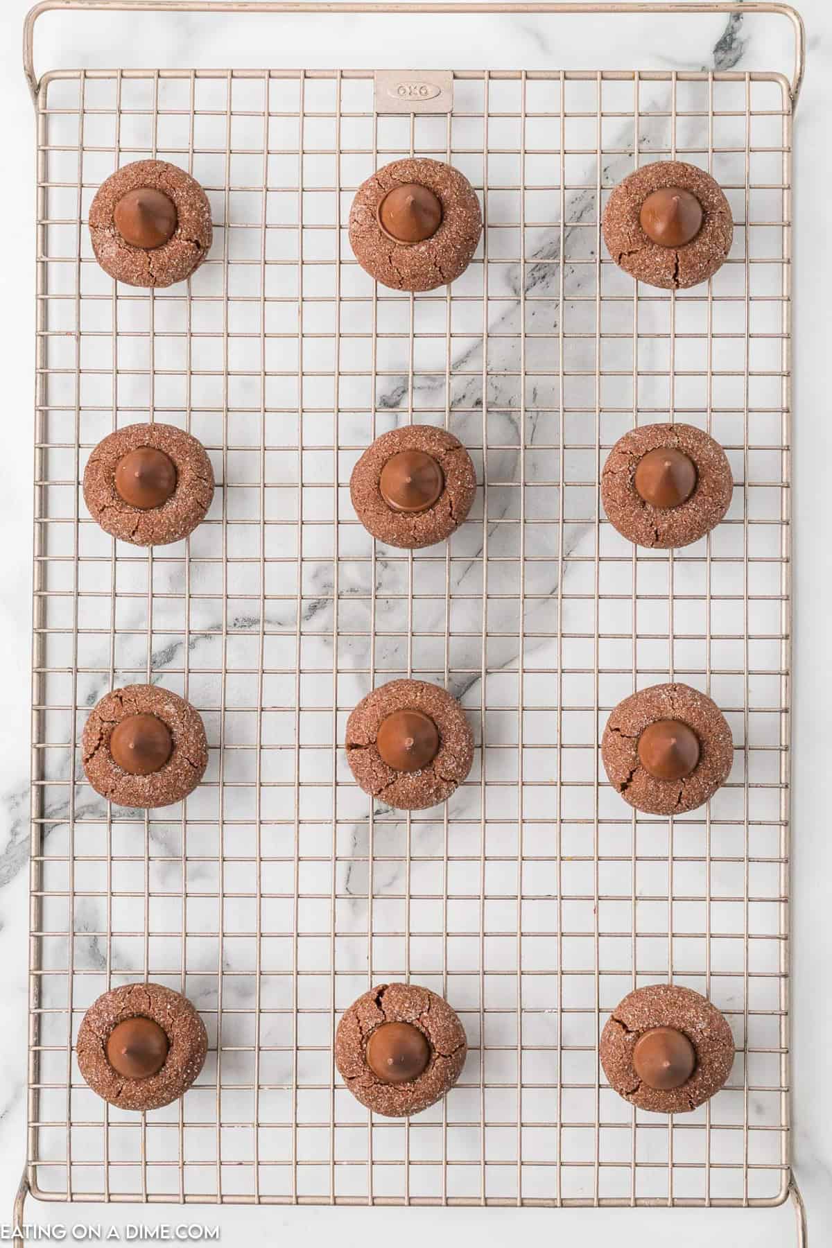 A cooling rack holds three rows of Chocolate Kiss Cookies, each featuring a round chocolate candy pressed into the center. They're arranged neatly in a grid pattern on the rack, which sits on a marble surface.