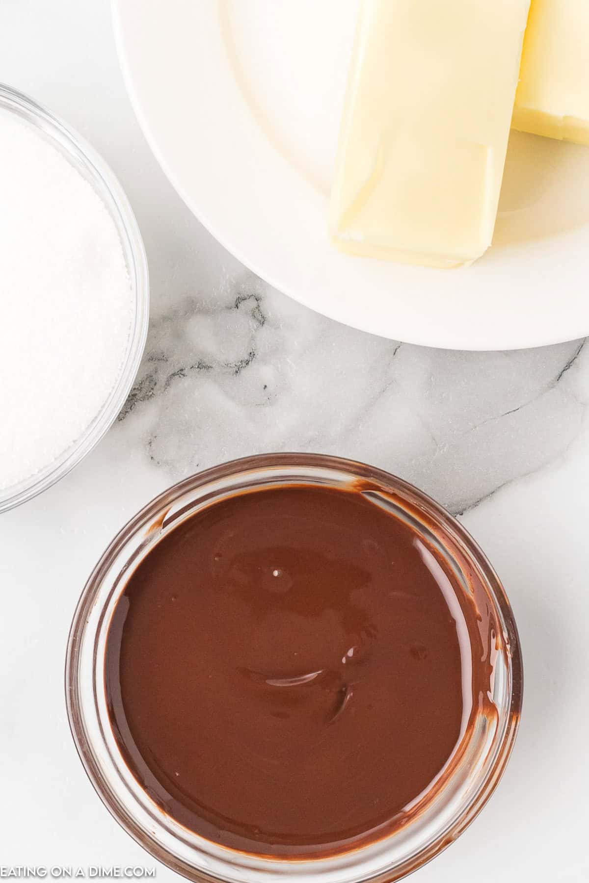 A bowl of melted dark chocolate, perfect for Chocolate Kiss Cookies, sits on a marble countertop. Next to it are a plate with two sticks of butter and a bowl of granulated sugar, ready to transform into decadent treats.