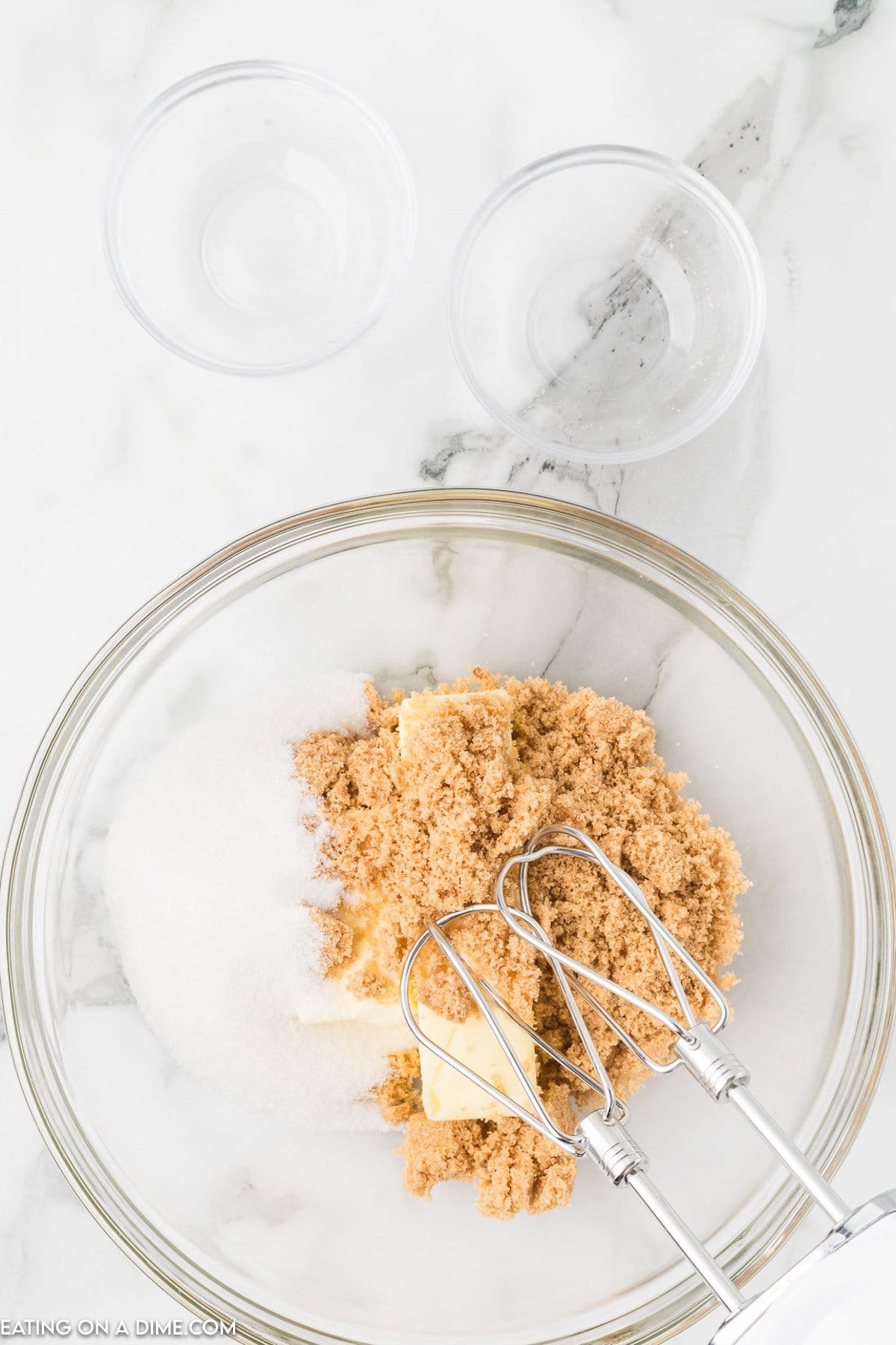 A glass bowl filled with brown sugar, white sugar, and butter rests on a marble countertop, ready to create delightful Chocolate Kiss Cookies. A handheld mixer with beaters sits poised inside the bowl. Two small empty glass bowls are placed above it, waiting for their turn in the baking process.