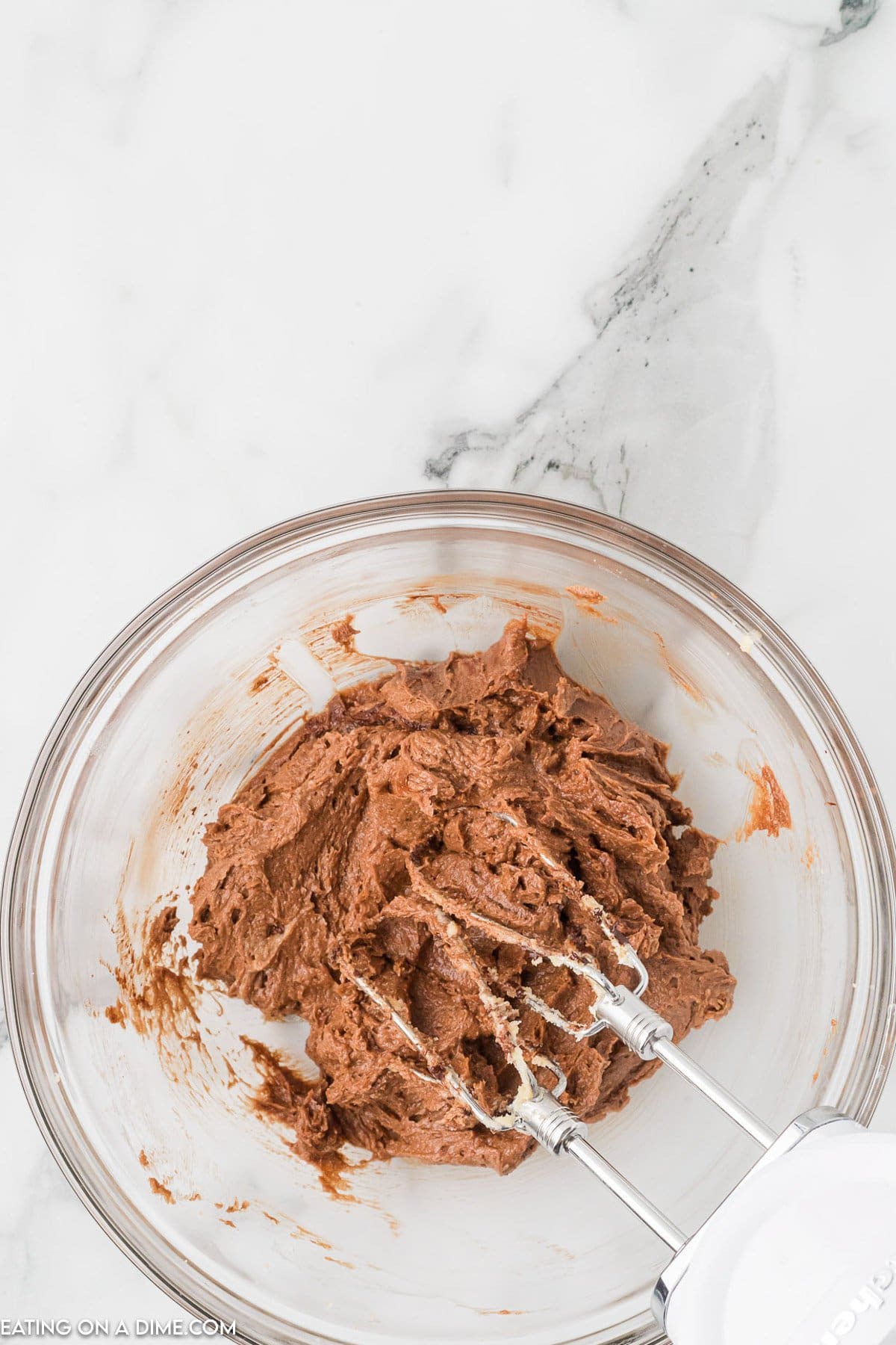 A glass bowl filled with chocolate cookie dough rests on a marble countertop, ready to transform into delectable chocolate kiss cookies. An electric hand mixer with two beaters sits in the dough, hinting at the recent mixing that promises a sweet indulgence.