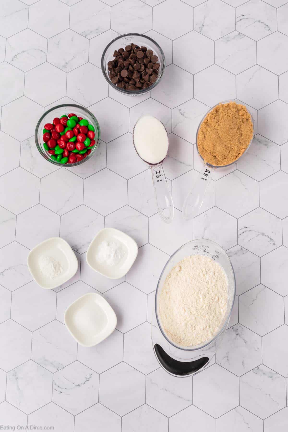 Ingredients for Christmas cookies in a jar are artfully arranged on a hexagonal tile surface. This festive assortment includes chocolate chips, red and green candy-coated chocolates, brown sugar, white sugar, baking powder, baking soda, salt, and flour—all neatly organized in bowls and measuring cups.