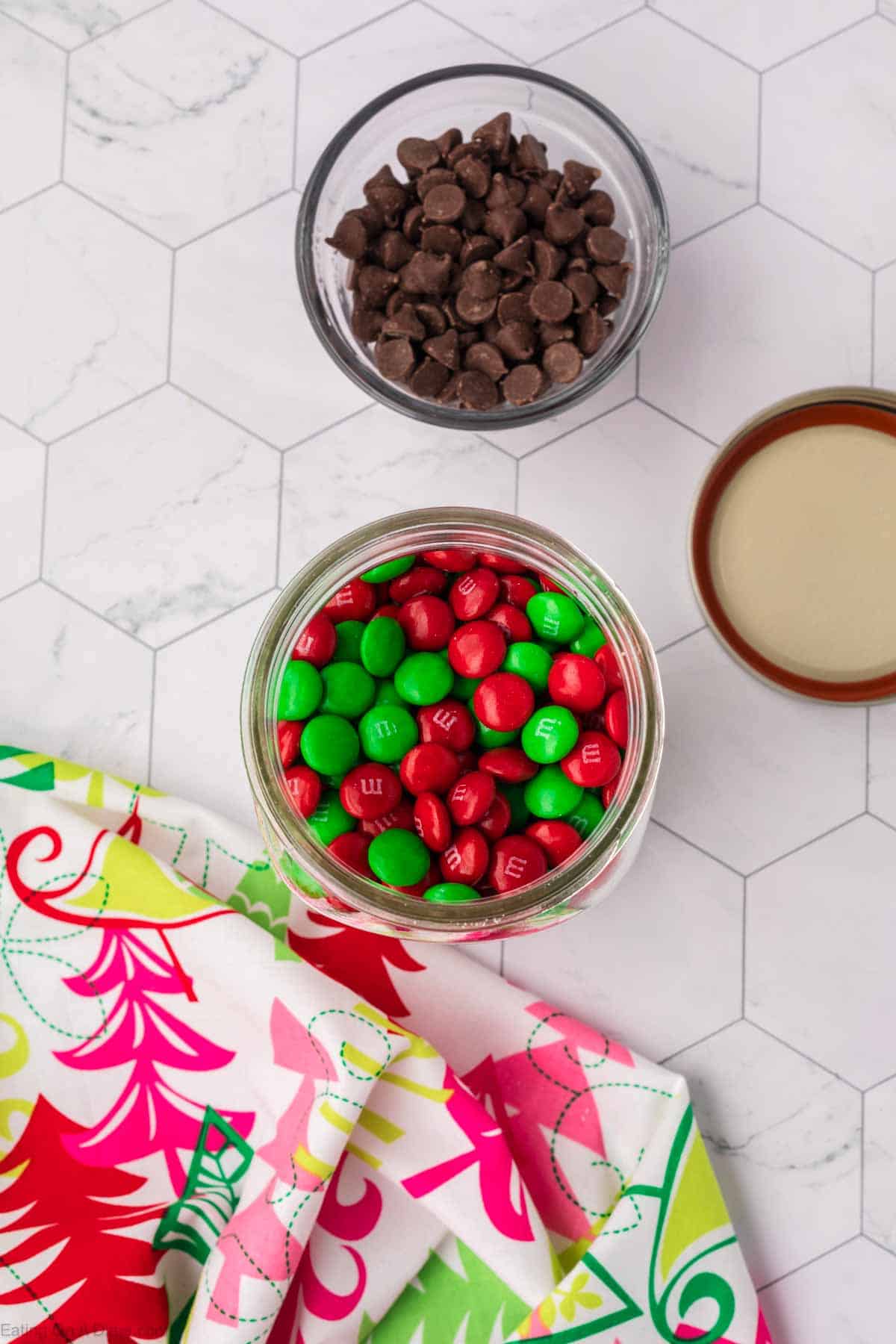 A jar filled with green and red M&Ms sits on a hexagonal patterned surface, evoking the festive vibes of Christmas cookies in a jar. Nearby is a bowl of chocolate chips and a colorful fabric with a holiday theme, while a wooden lid rests off to the side.