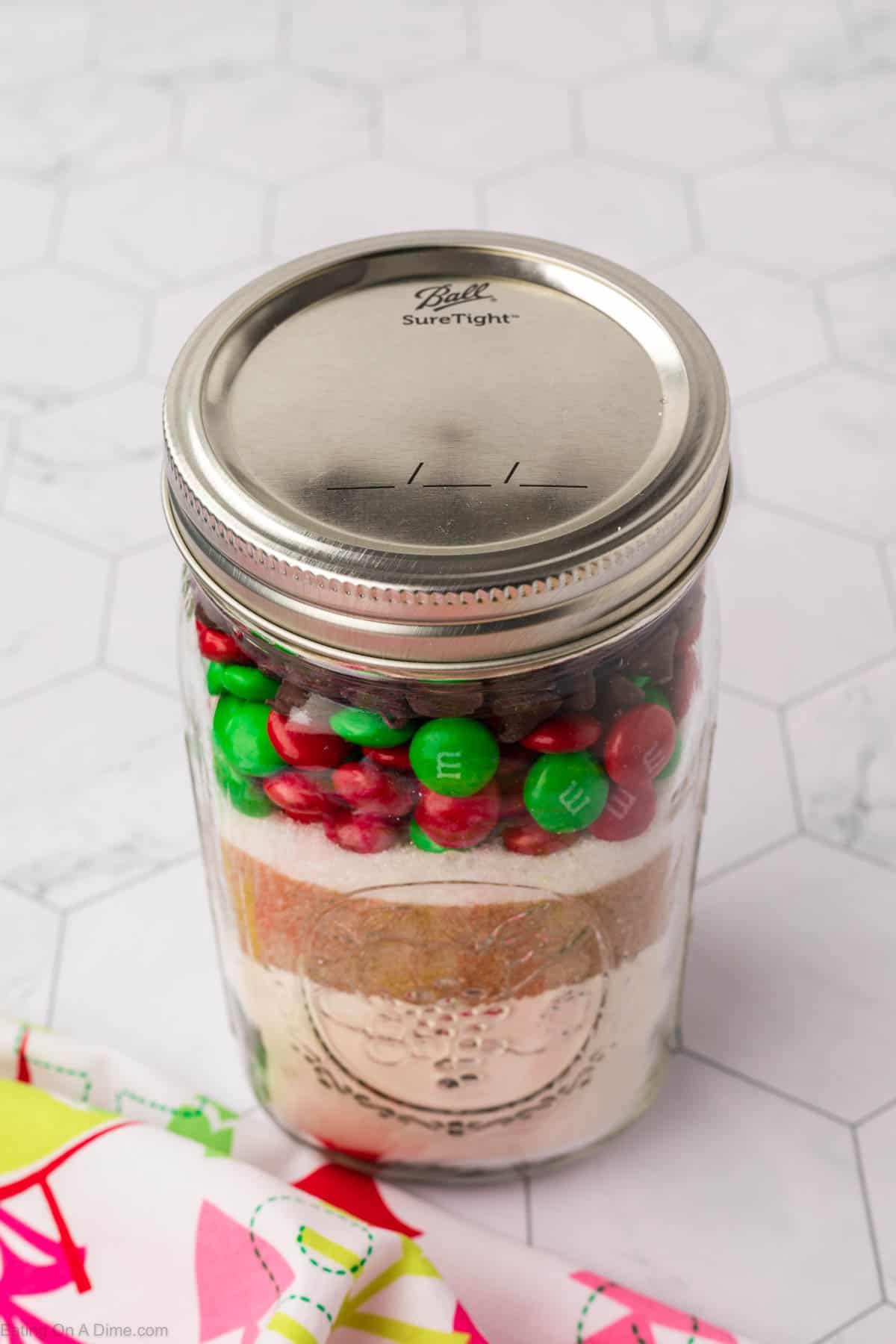 A glass jar with a silver lid is filled with layers of white flour, brown sugar, and colorful candy-coated chocolates, creating a festive scene reminiscent of Christmas Cookies in a Jar. It rests on a white surface with a light hexagonal pattern, next to a brightly colored cloth.