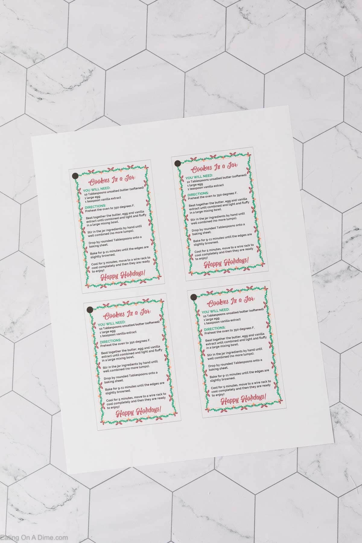 Four festive recipe cards titled "Christmas Cookies in a Jar" are arranged on a marble hexagon-patterned surface. Each card lists ingredients and instructions for holiday cookies, framed with a decorative red and green border.