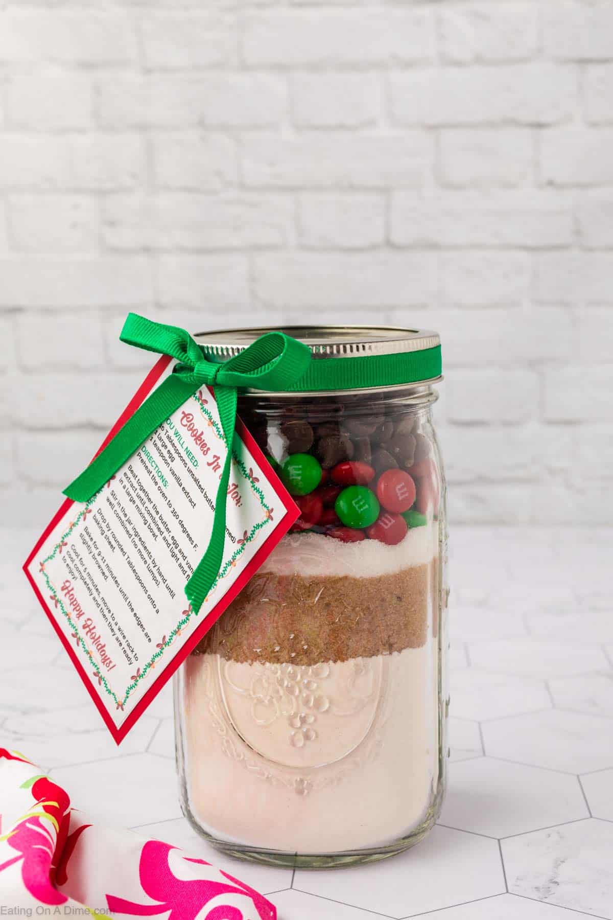 A festive jar of dry ingredients for Christmas Cookies in a Jar, topped with chocolate chips and red and green candy-coated chocolates. A green ribbon with a tag wraps around the lid, featuring baking instructions. The background is a white brick wall.