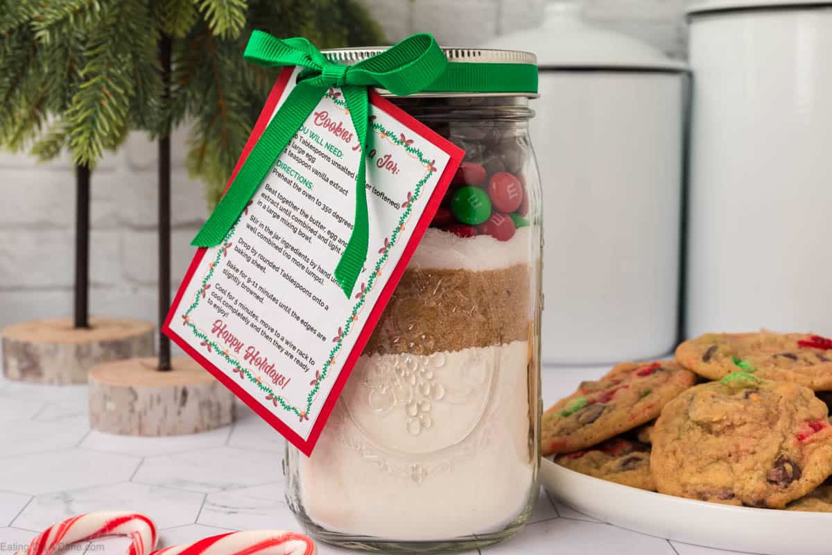 A jar of Christmas Cookies in a Jar, with ingredients layered inside, is adorned with a green bow and a red-bordered recipe card. It sits on a kitchen countertop beside a small pine tree and a plate of cookies, surrounded by scattered candy canes.