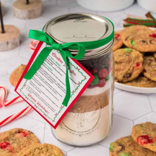 A mason jar filled with Christmas cookies in a jar ingredients, topped with a green ribbon and a recipe card. In the background, there are cookies, candy canes, and festive decorations adorning a white hexagonal tablecloth.