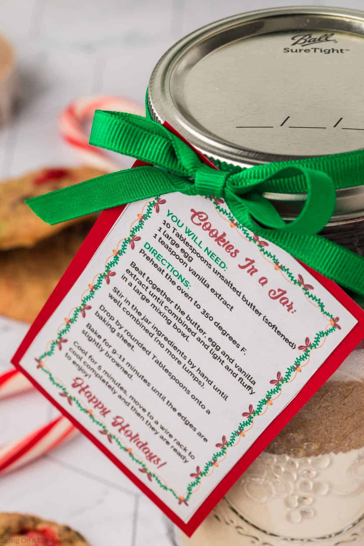 A jar adorned with red and green ribbons, showcasing a festive recipe card for "Christmas Cookies in a Jar." The card details ingredients and directions, framed by holiday decorations. Blurred cookies and candy canes add to the seasonal ambiance.