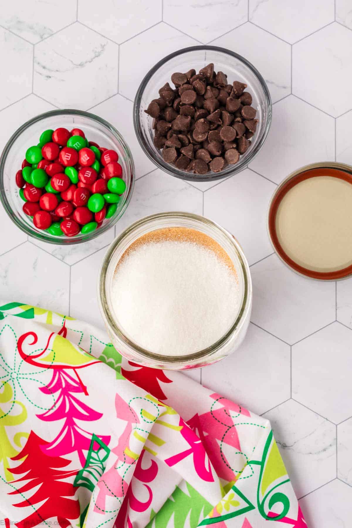 Overhead view of baking ingredients for festive Christmas cookies in a jar on a hexagon-tiled surface: a jar of sugar, a bowl of chocolate chips, red and green candies, and a brown-rimmed bowl. A colorful cloth with cheerful patterns lies nearby.