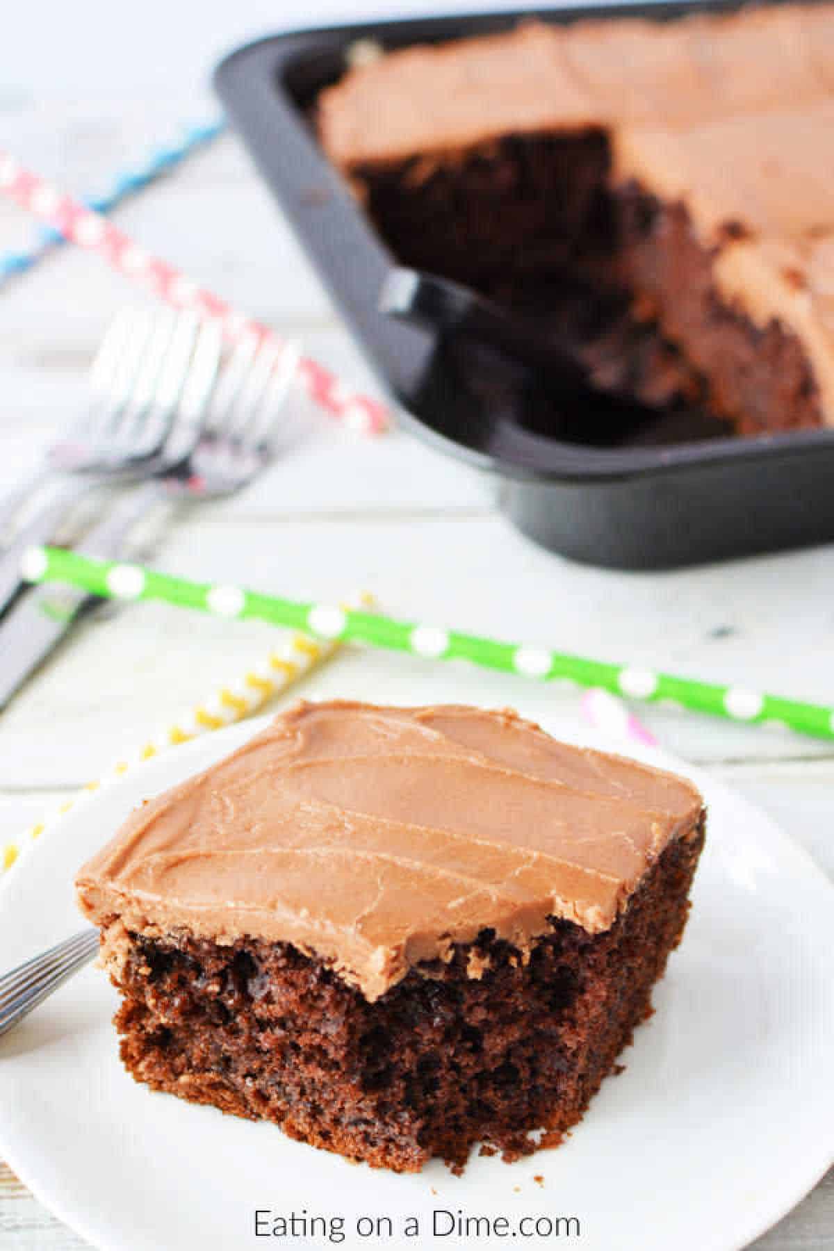 A slice of cola cake with frosting rests on a white plate beside a fork. In the background, a baking pan holds more cake, surrounded by colorful striped straws and silver forks on a light wooden surface.
.