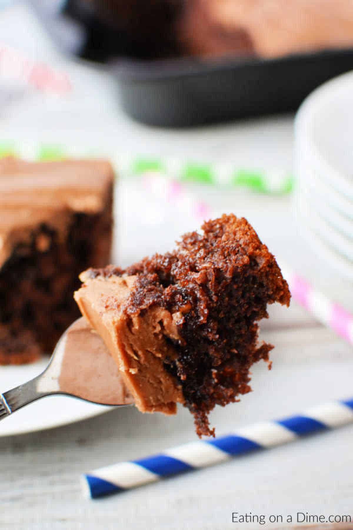 Close-up of a fork holding a bite of moist cola-infused chocolate cake with rich frosting. The background features a pan with more cake and colorful striped straws.