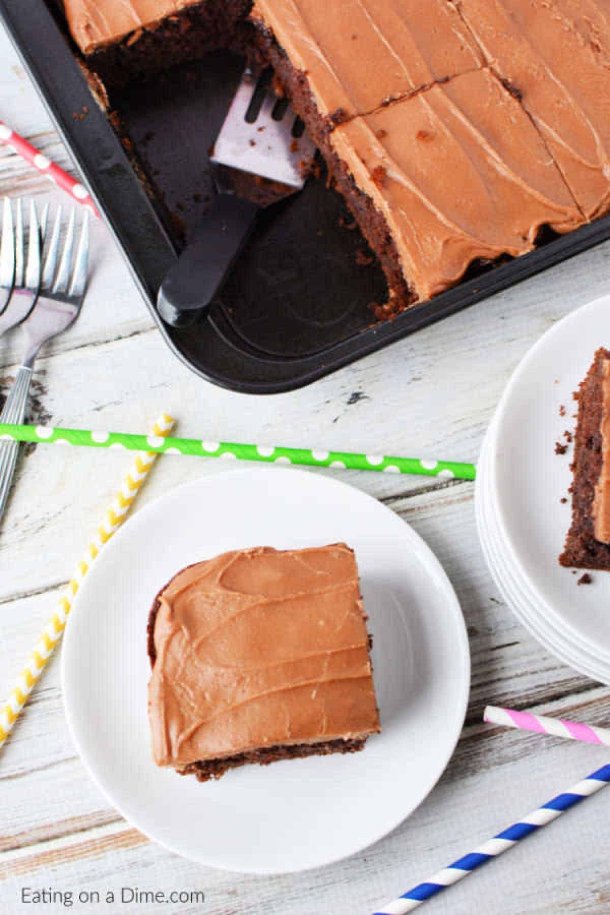 A frosted chocolate cola cake in a baking tray with a spatula. A piece is served on a white plate next to colorful striped straws and a stack of forks on a white wooden table.