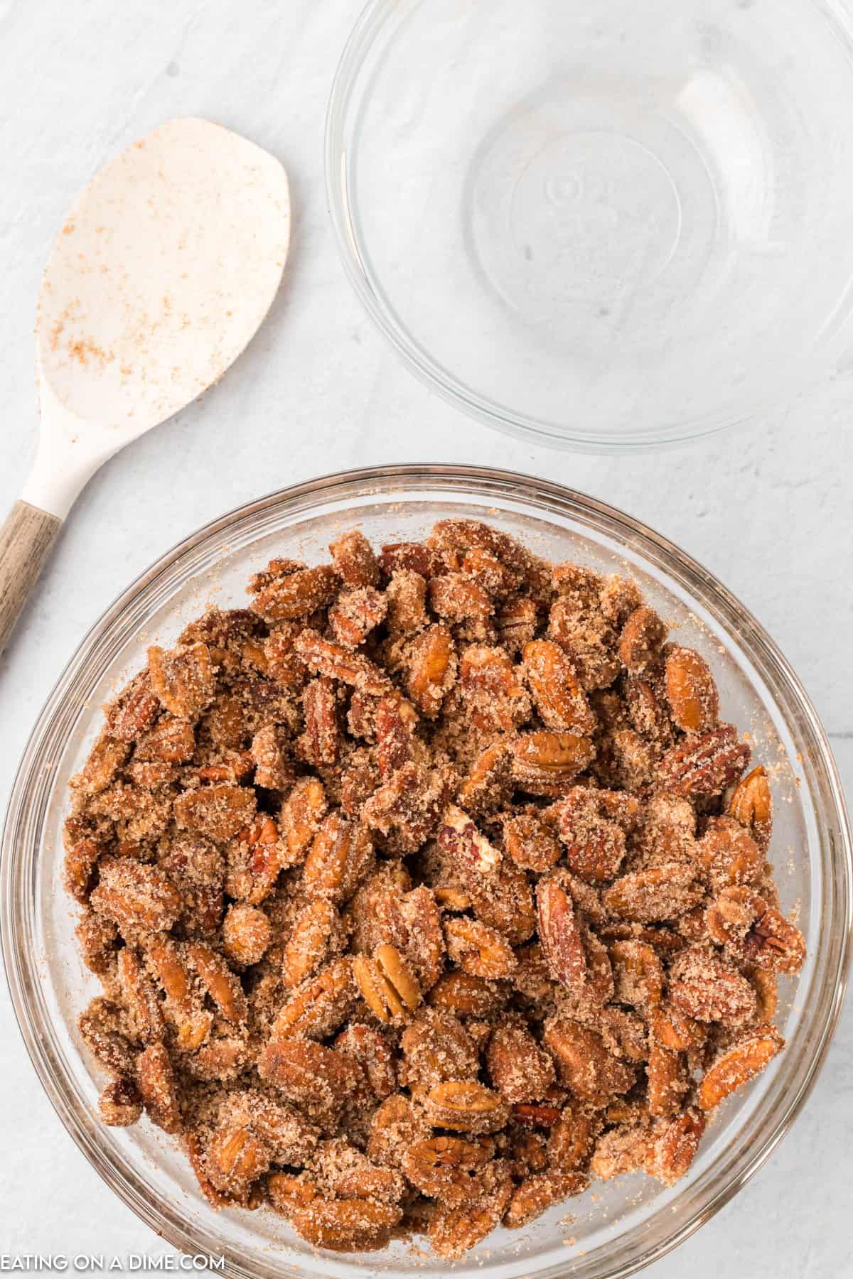 A glass bowl filled with slow cooker candied pecans, placed on a white surface. A wooden spoon with a white, slightly stained scoop is situated next to the bowl, and an empty glass bowl is visible in the background.