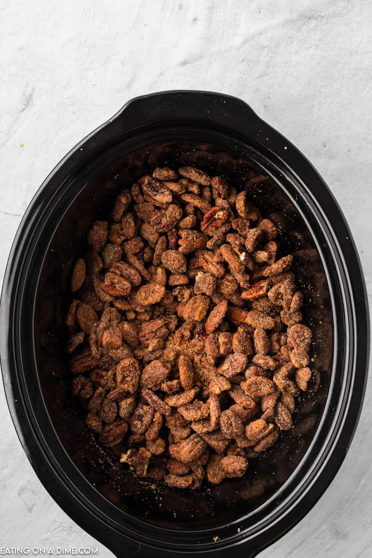 Top view of a slow cooker filled with seasoned and roasted nuts, including delicious Slow Cooker Candied Pecans. The nuts are coated in a brown spice mixture, evenly distributed inside the cooker. The background is a light grey surface. A small part of the slow cooker lid is visible at the top left corner.