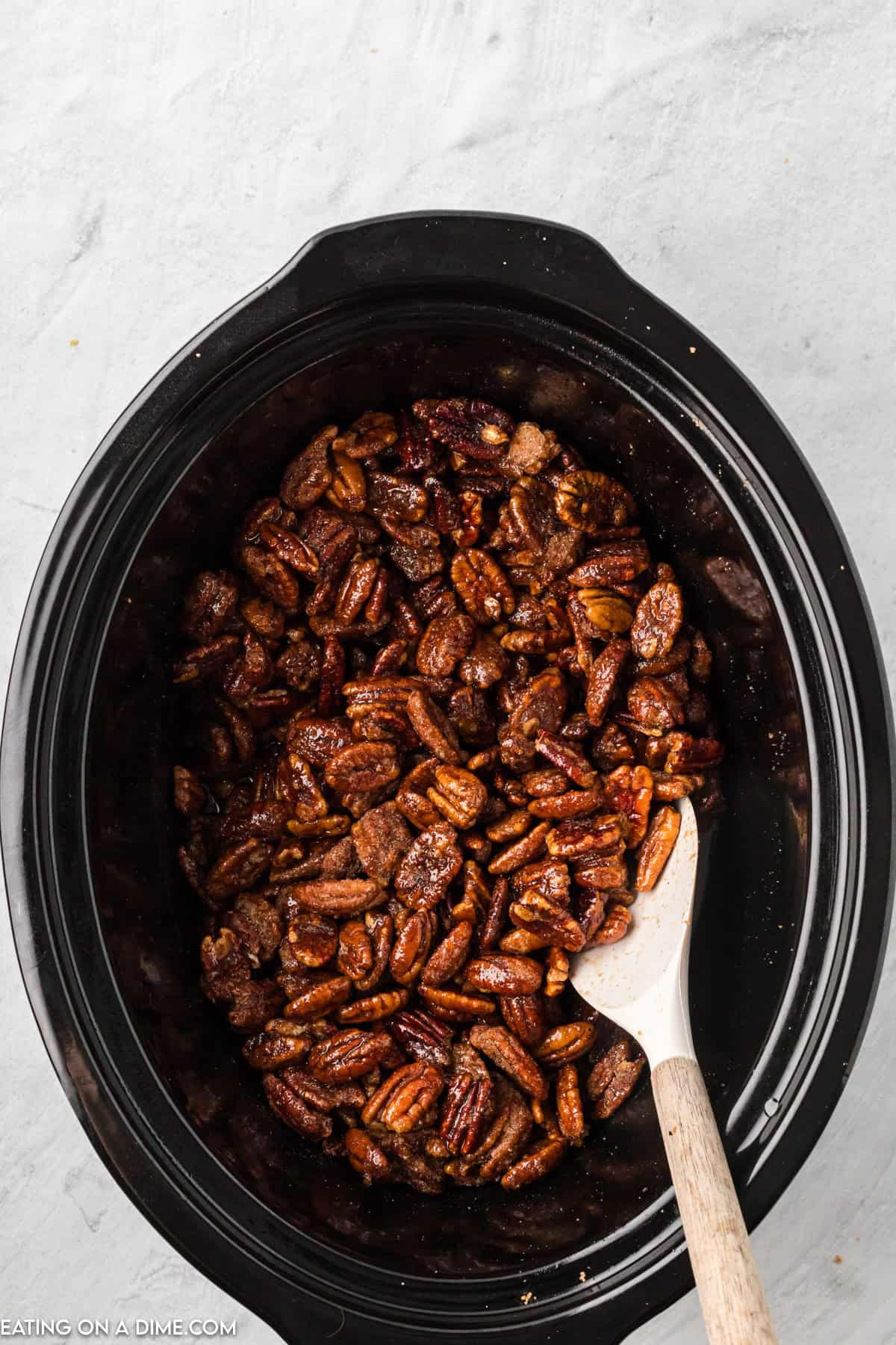 A slow cooker filled with Slow Cooker Candied Pecans is placed on a light gray surface. A wooden spoon with a white handle rests inside the slow cooker, partially submerged in the deliciously glazed pecans.