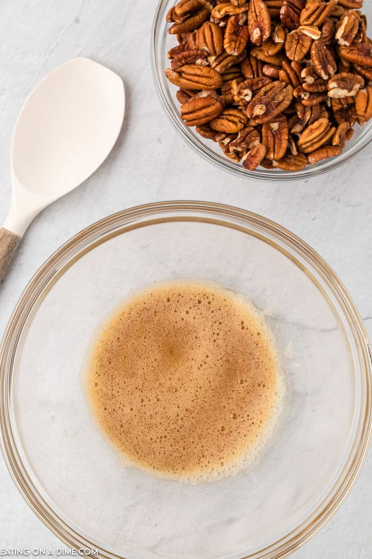 A glass bowl containing a frothy brown liquid sits on a white countertop. Next to it, a white spoon with a wooden handle rests near another glass bowl filled with slow cooker candied pecans.