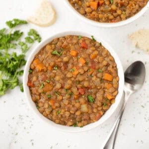 A bowl of slow cooker lentil soup filled with lentils, diced carrots, and herbs sits invitingly. A spoon rests beside the bowl, while fresh parsley and pieces of bread adorn the white surface in the background.