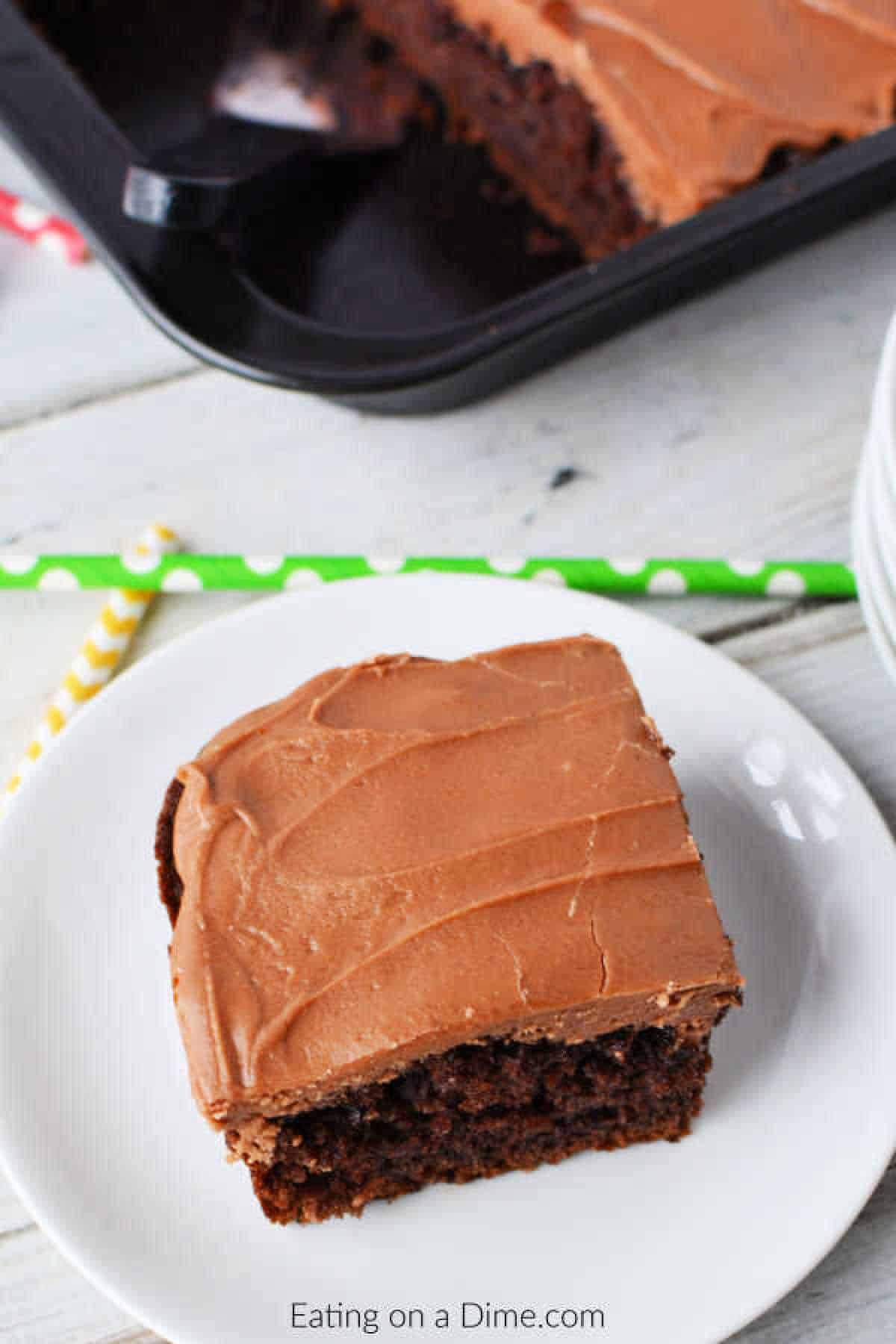 A slice of cola chocolate cake with smooth frosting rests on a white plate. In the background, a baking tray holds more of this delightful treat. Yellow and green striped straws are placed beside the plate on a wooden table, adding a pop of color to the scene.