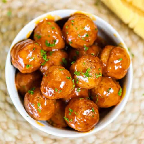 A bowl filled with shiny crock pot honey buffalo chicken meatballs topped with chopped parsley sits on a woven surface. The meatballs glisten with a rich sauce, and a small portion of yellow fabric is visible at the edge of the image.