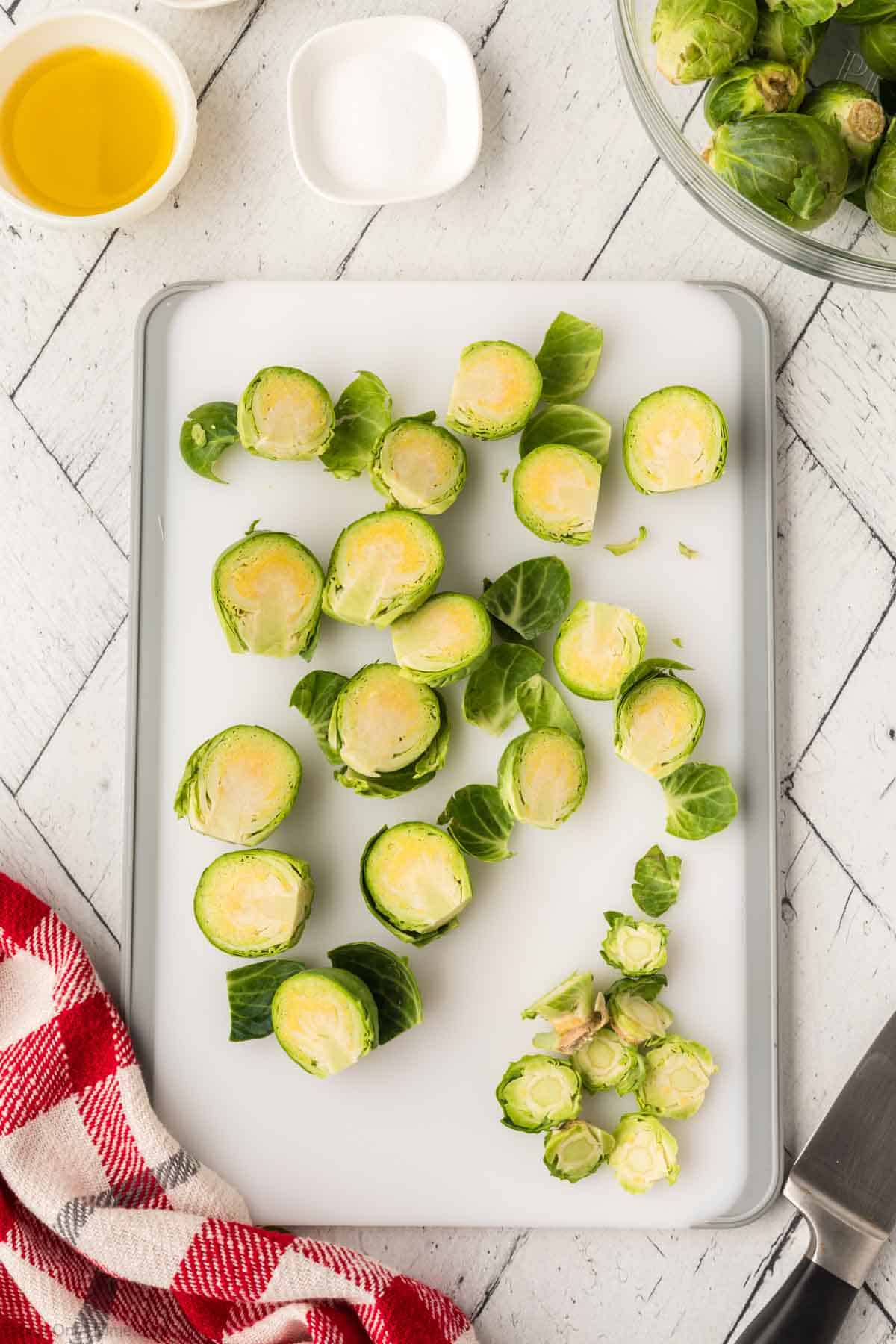 Cut Brussel Sprouts in half on a cutting board with small bowls of oil and salt on the side