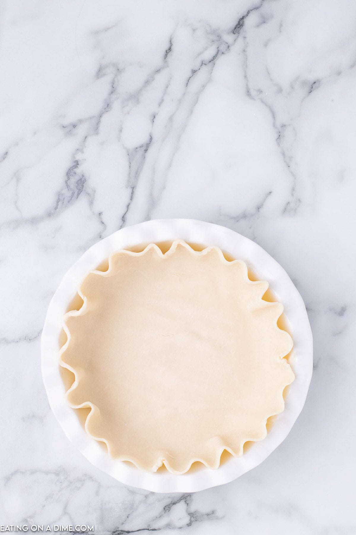 A pie crust with crimped edges is placed in a white pie dish on a marbled countertop, ready to transform into a delightful cinnamon pie recipe.