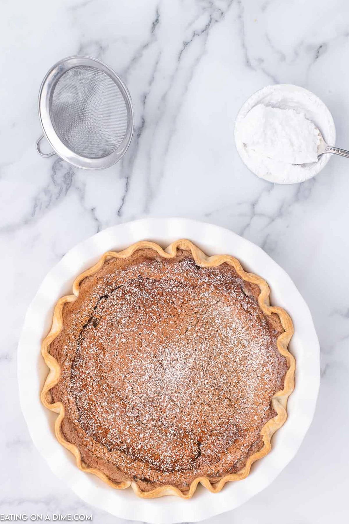 A baked chocolate chess pie in a white pie dish, dusted with powdered sugar, offers an inviting twist on the classic cinnamon pie recipe. A metal sieve and a small bowl of powdered sugar with a spoon rest beside it on a marble surface.