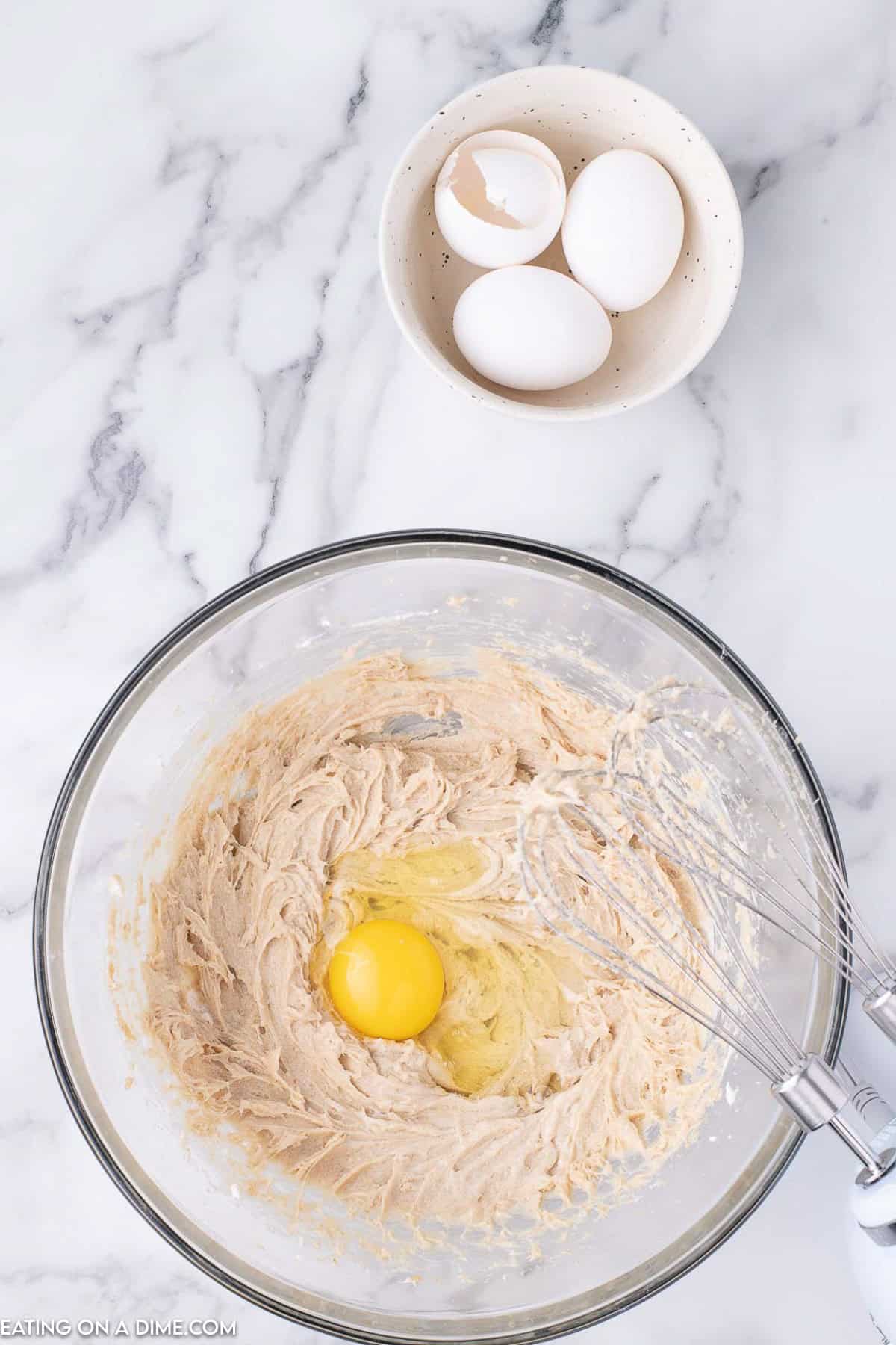 A whisk in a bowl of creamy mixture, hinting at a cinnamon pie recipe, with a raw egg on top sits on a marble countertop. Nearby, a bowl holds three whole eggs, one partially cracked.