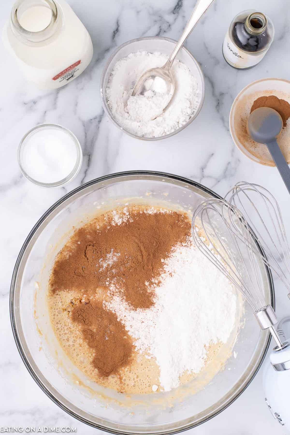 On a marble countertop, a mixing bowl holds a blend of ingredients for the perfect cinnamon pie recipe. Alongside rests a whisk, with a bottle of vanilla extract, a jar of milk, and small bowls of flour and sugar neatly surrounding the scene.