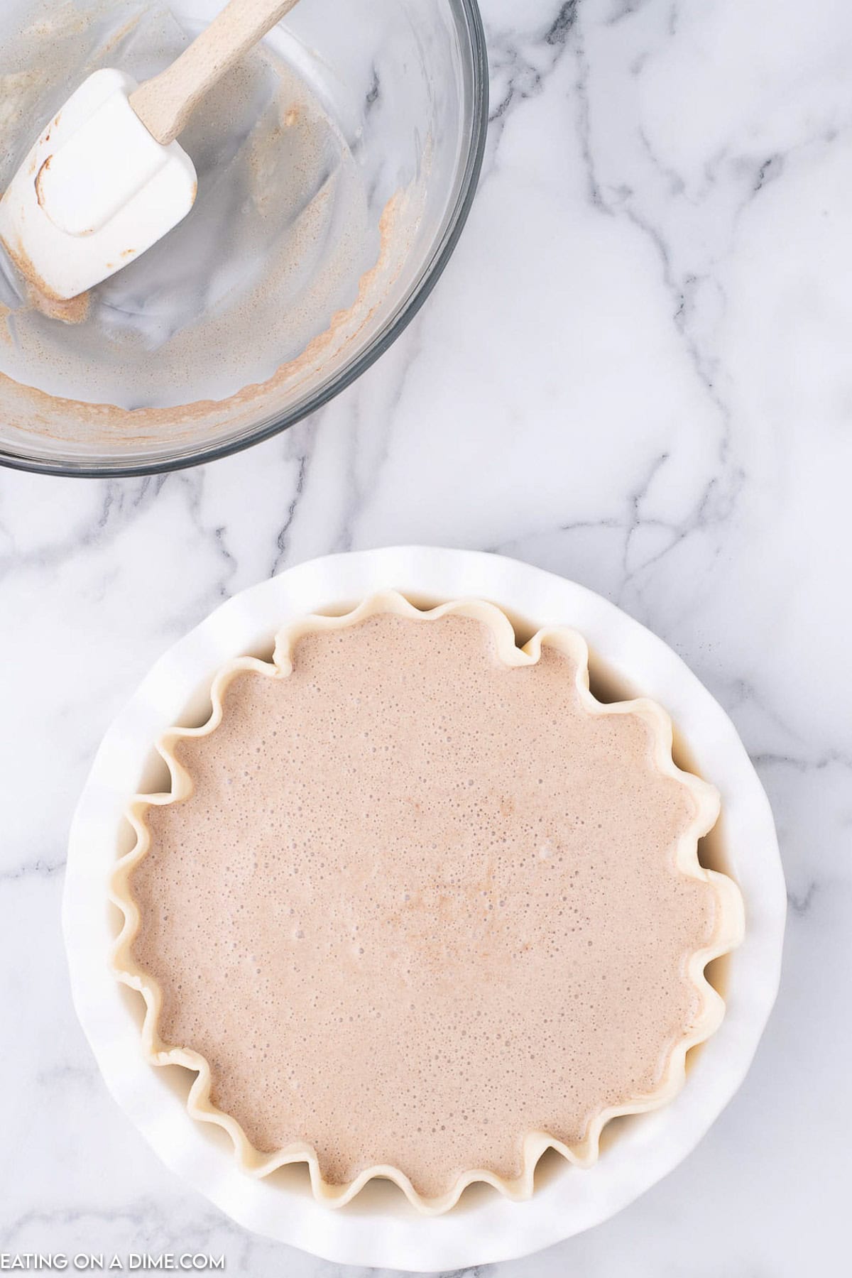 A pie crust filled with light brown cinnamon pie batter sits on a marble surface. A spatula rests in a glass mixing bowl with remnants of the batter nearby. The pie, part of the perfect cinnamon pie recipe, has a wavy edge.